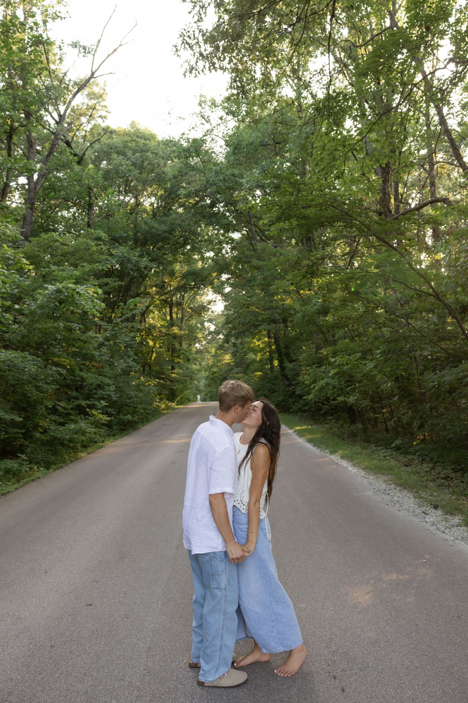 A couple shares a romantic moment on a secluded forest road surrounded by green trees.