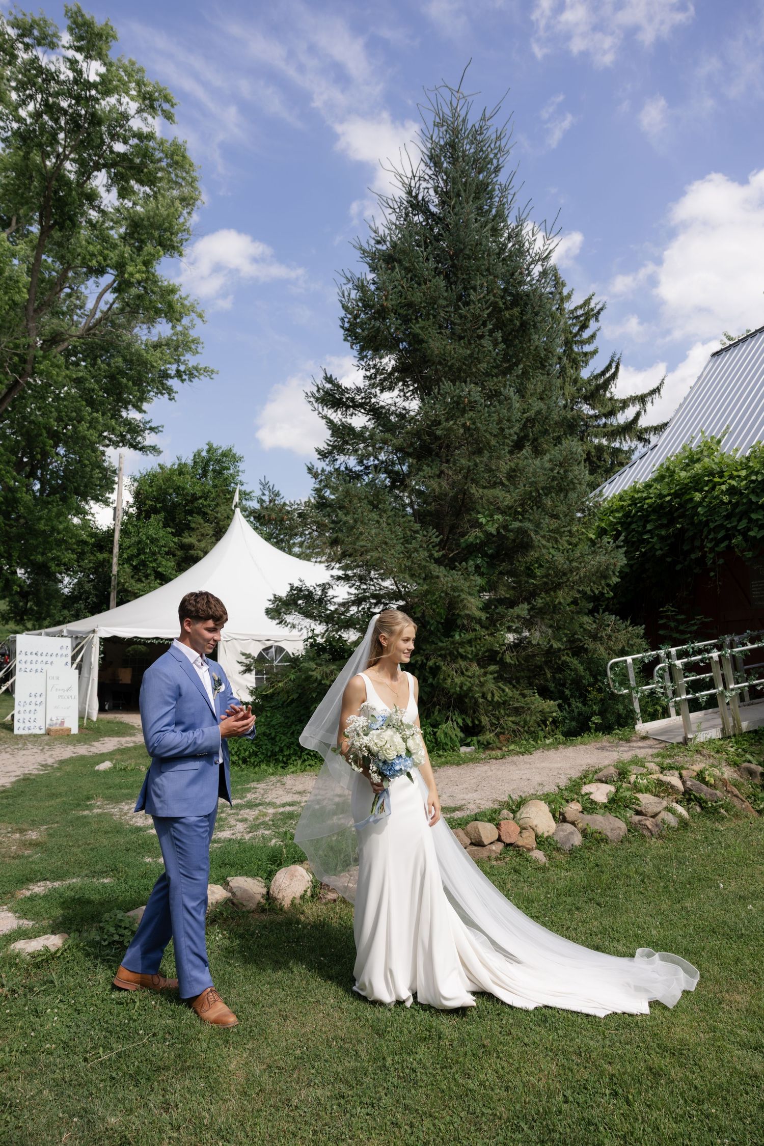 Wedding couple walking on grassy hill with white tent and evergreen trees in background.