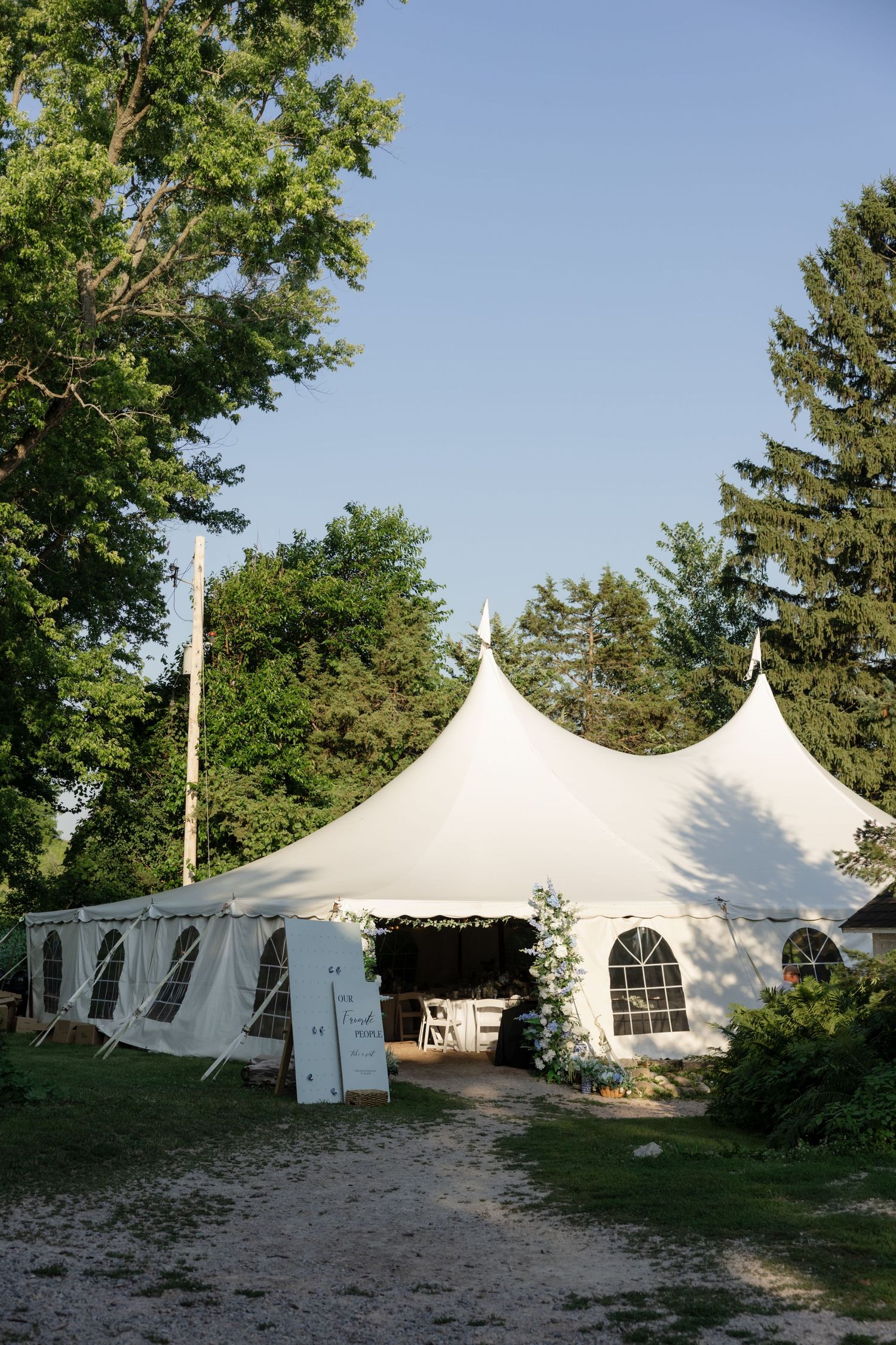 A white wedding tent with peaked tops stands elegantly among trees on a summer day.
