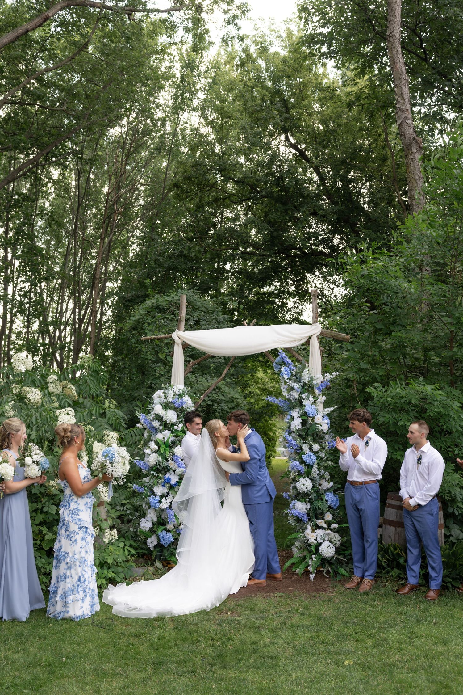 Wedding party stands together in a line against green trees and blue floral decor.