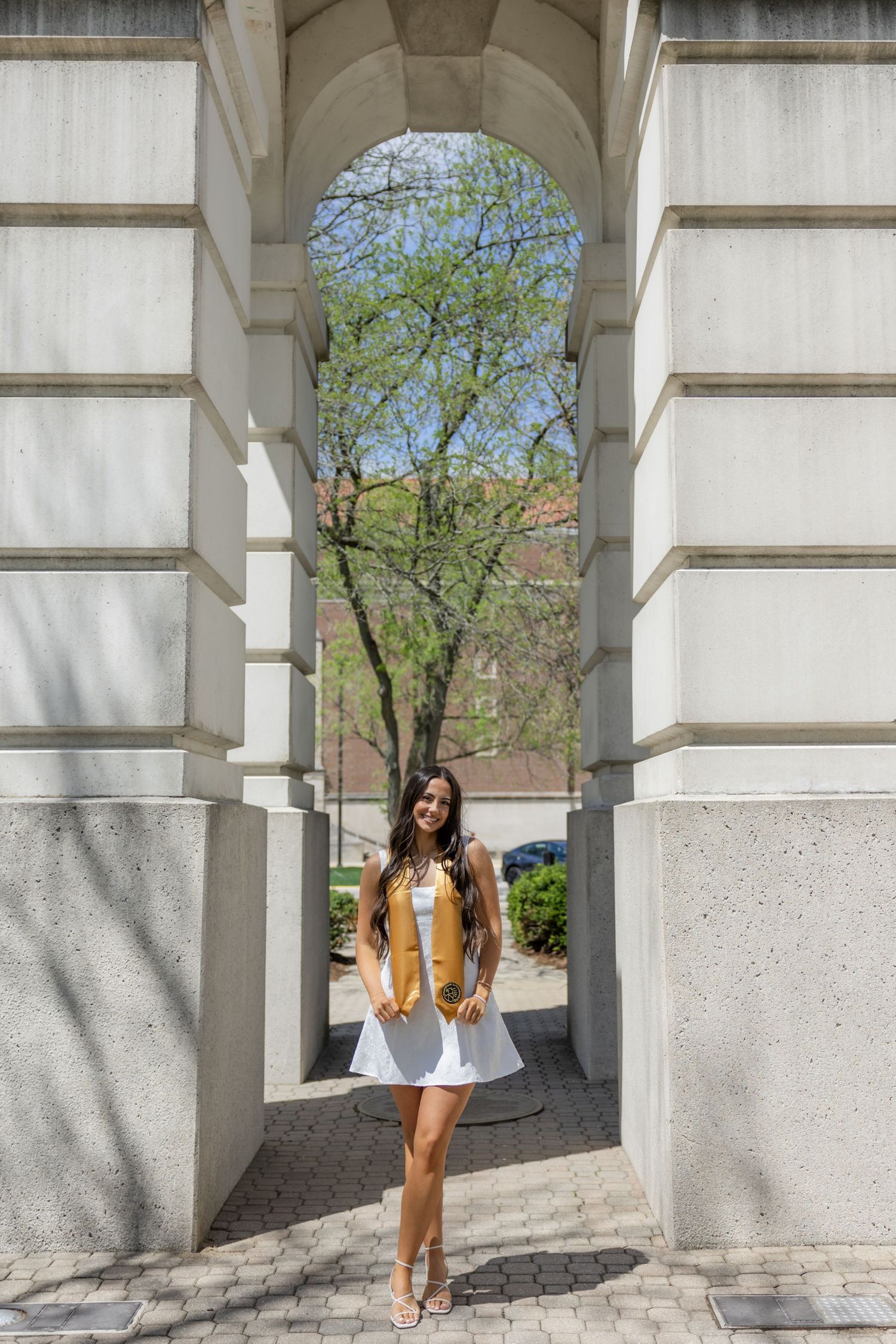 Graduate in white dress and gold sash stands under stone archway on sunny campus.