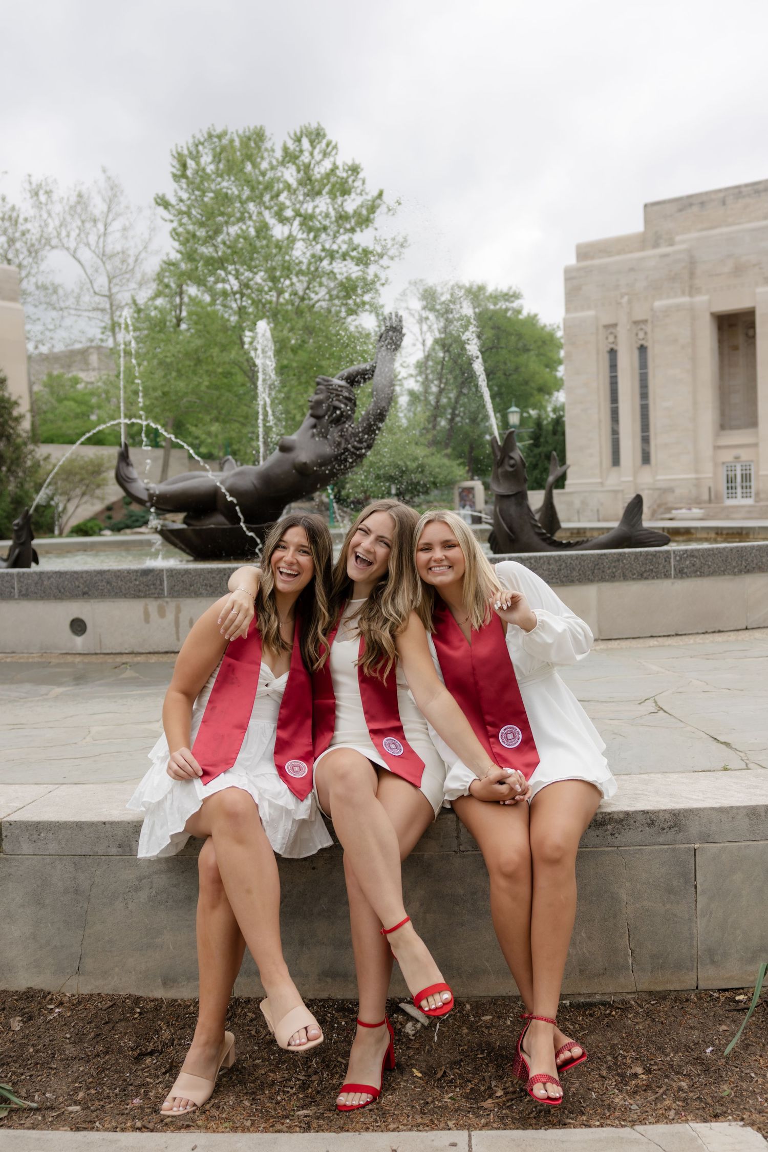 Three graduates in matching white dresses and red sashes pose together by a fountain statue.