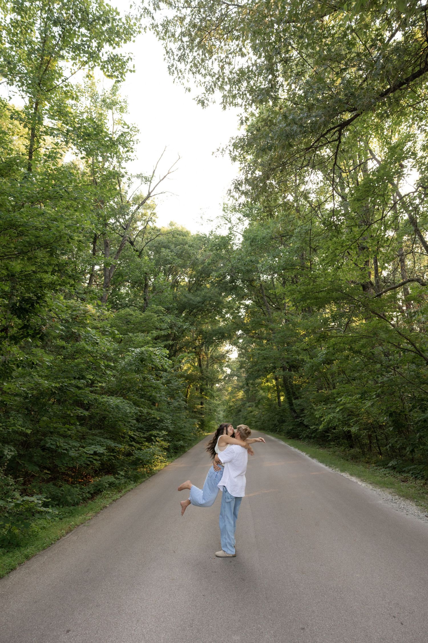 A couple embraces in the middle of a scenic tree-lined road on a summer afternoon.