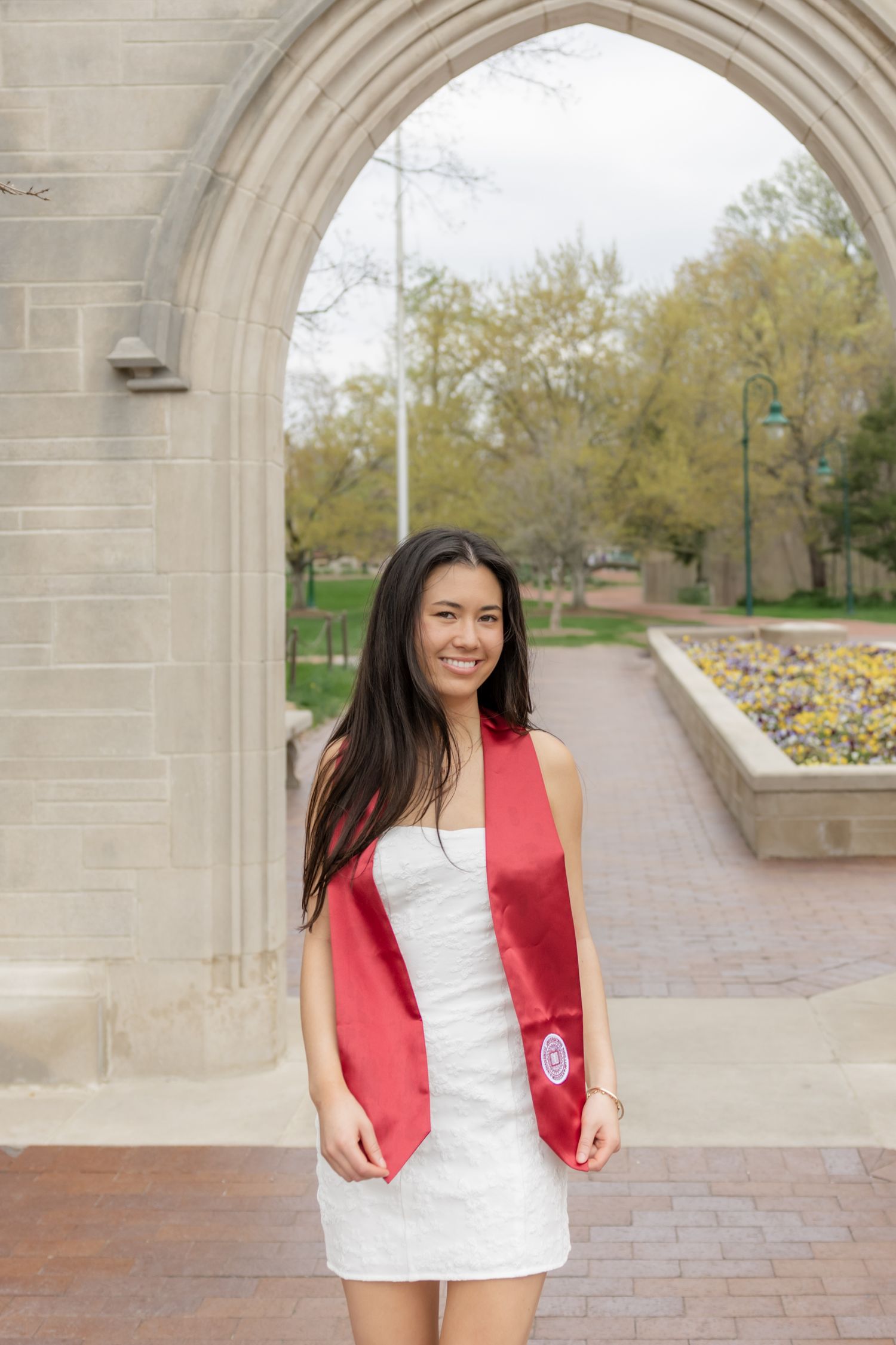A person in graduation attire stands smiling under a stone archway on a university campus.