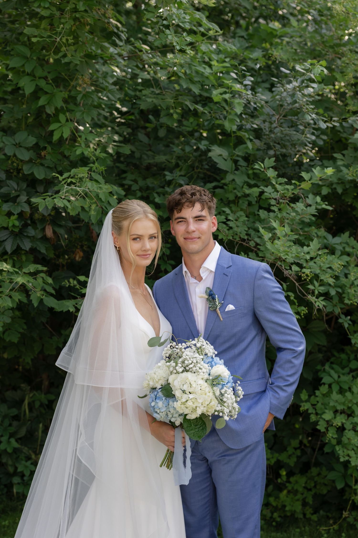 Wedding portraits against green foliage with blue suit and white floral bouquet.