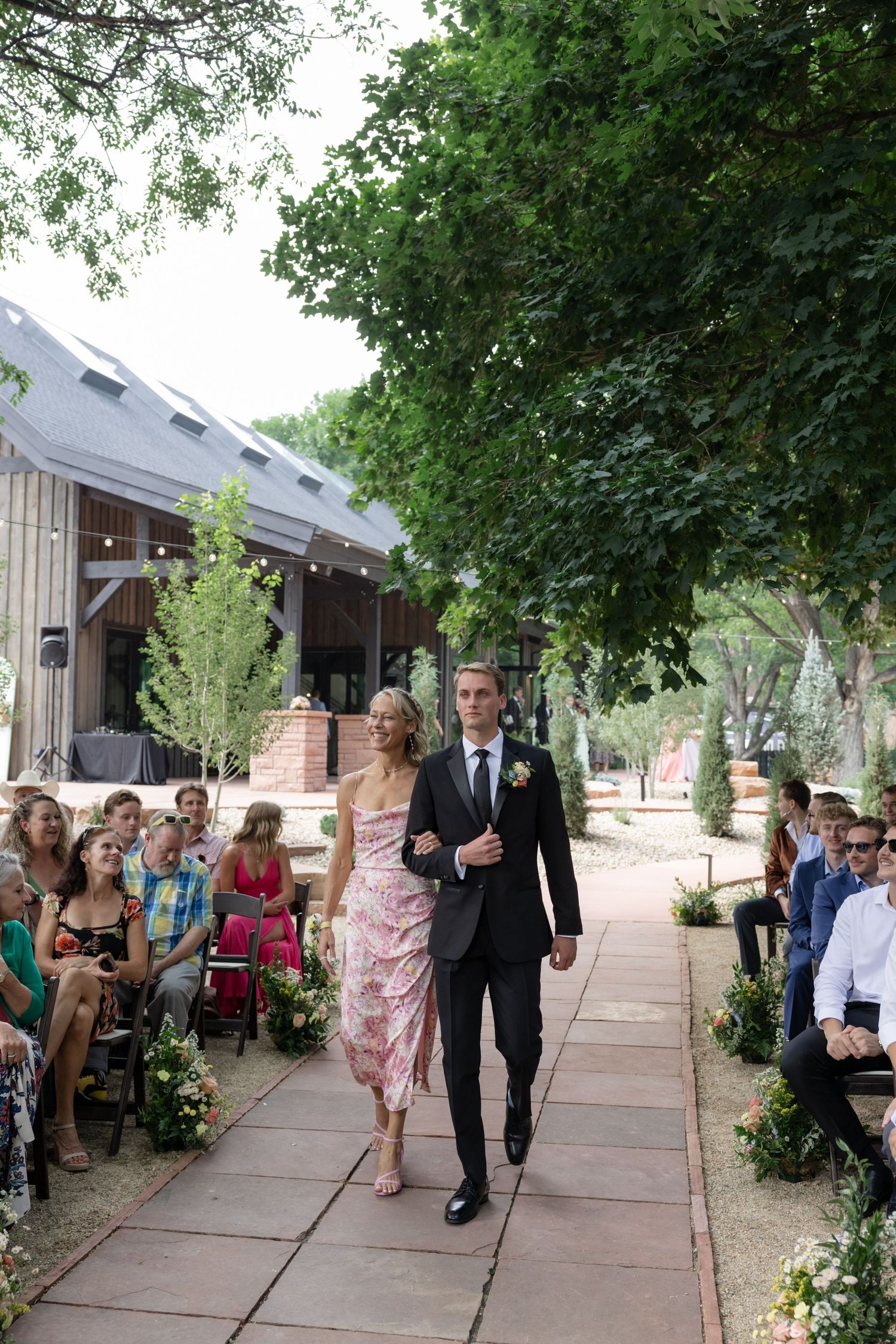 Wedding ceremony processional on a stone pathway lined with greenery and seated guests.
