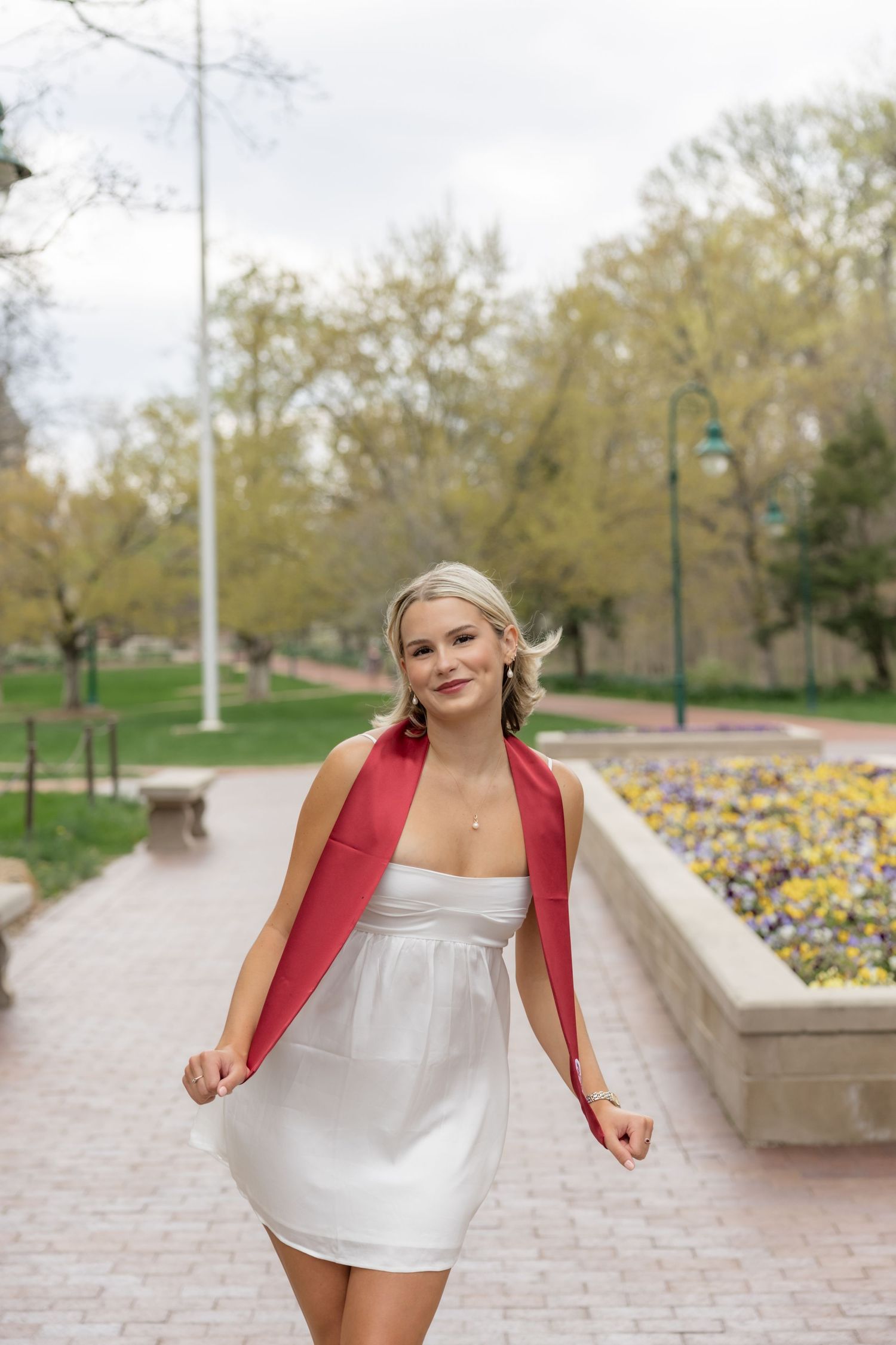 Person in white dress with red sash poses on outdoor campus pathway near yellow flowers.