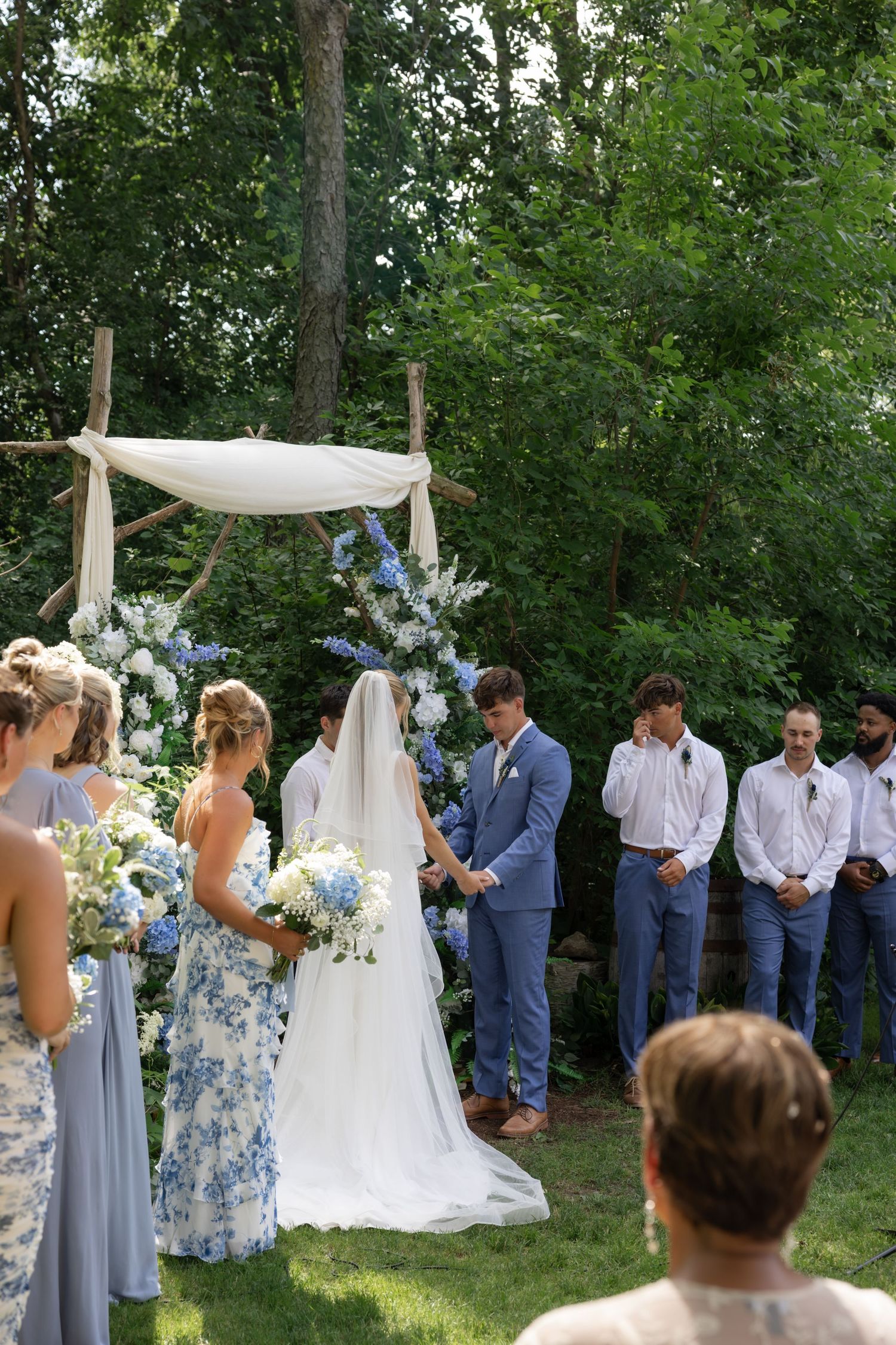 Wedding ceremony in progress with wedding party in blue suits and floral dresses under trees.