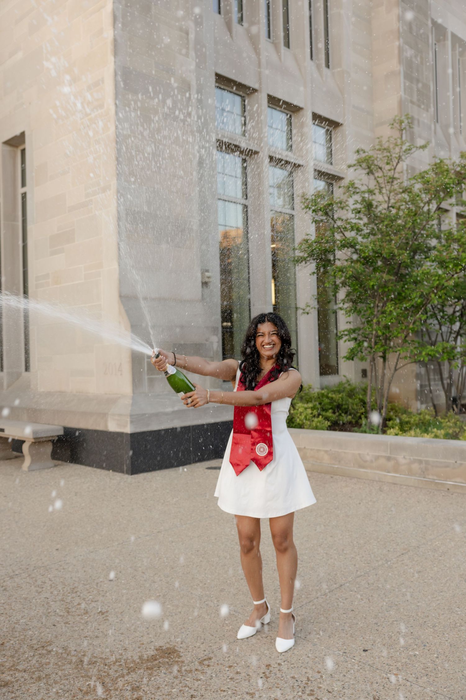 A person in graduation attire poses playfully outside a stone building.