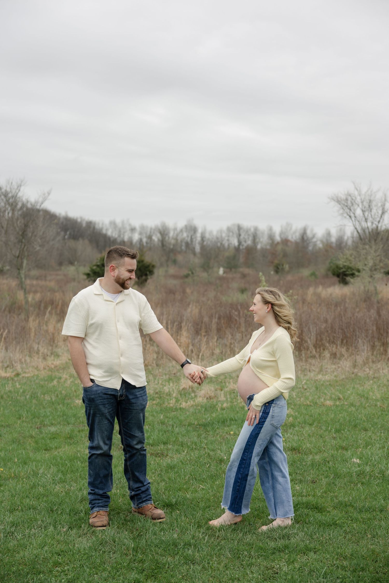 Full length portrait of couple holding hands in field during maternity photoshoot.