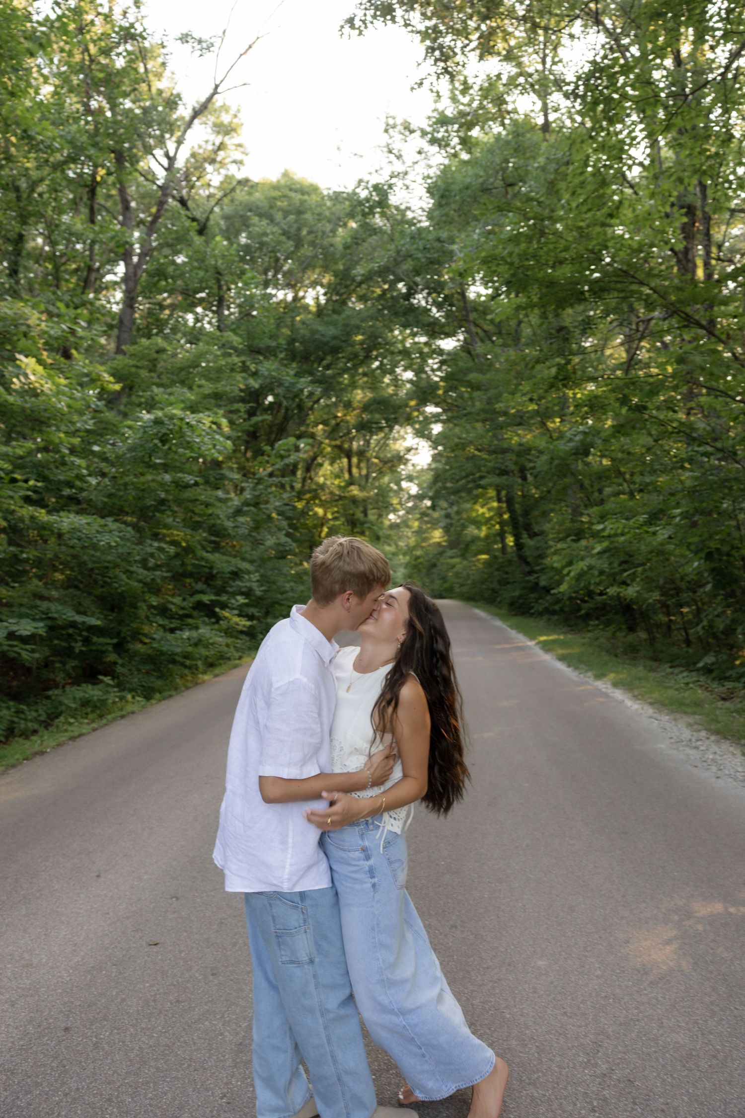 A romantic couple sharing a kiss on a tree-lined road during golden hour.