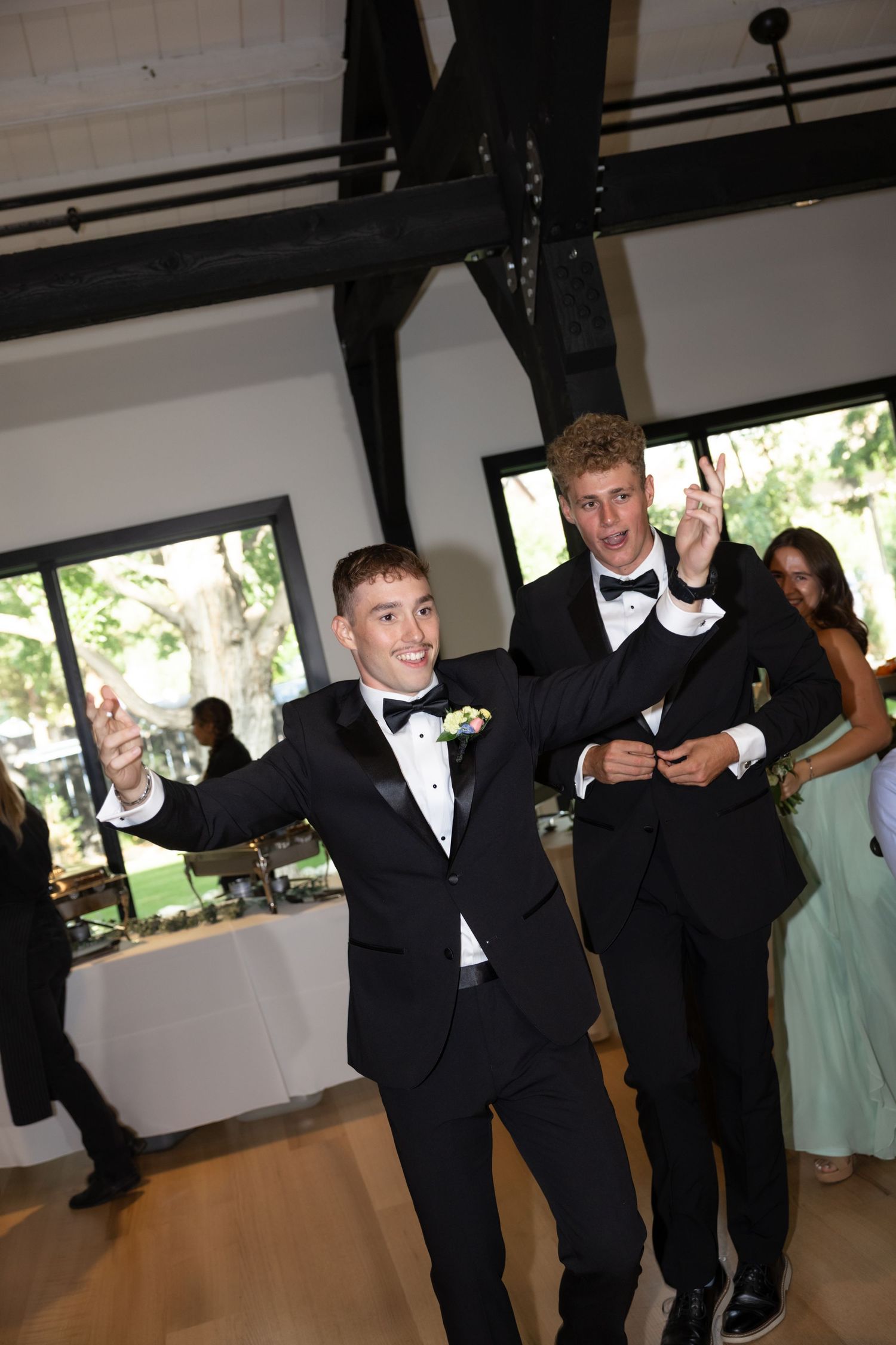Wedding party members in black tuxedos dance enthusiastically in an indoor reception venue.