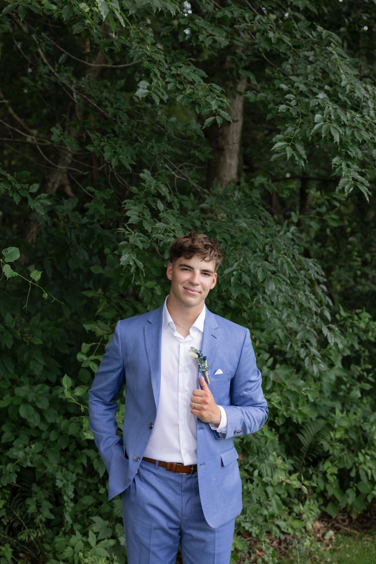 Person in a light blue suit and white shirt poses against a backdrop of green foliage.