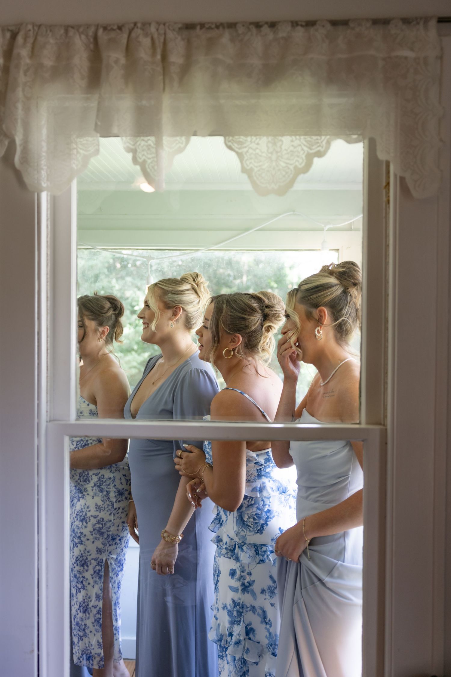Group wearing coordinated blue and floral dresses standing by window during wedding preparations.