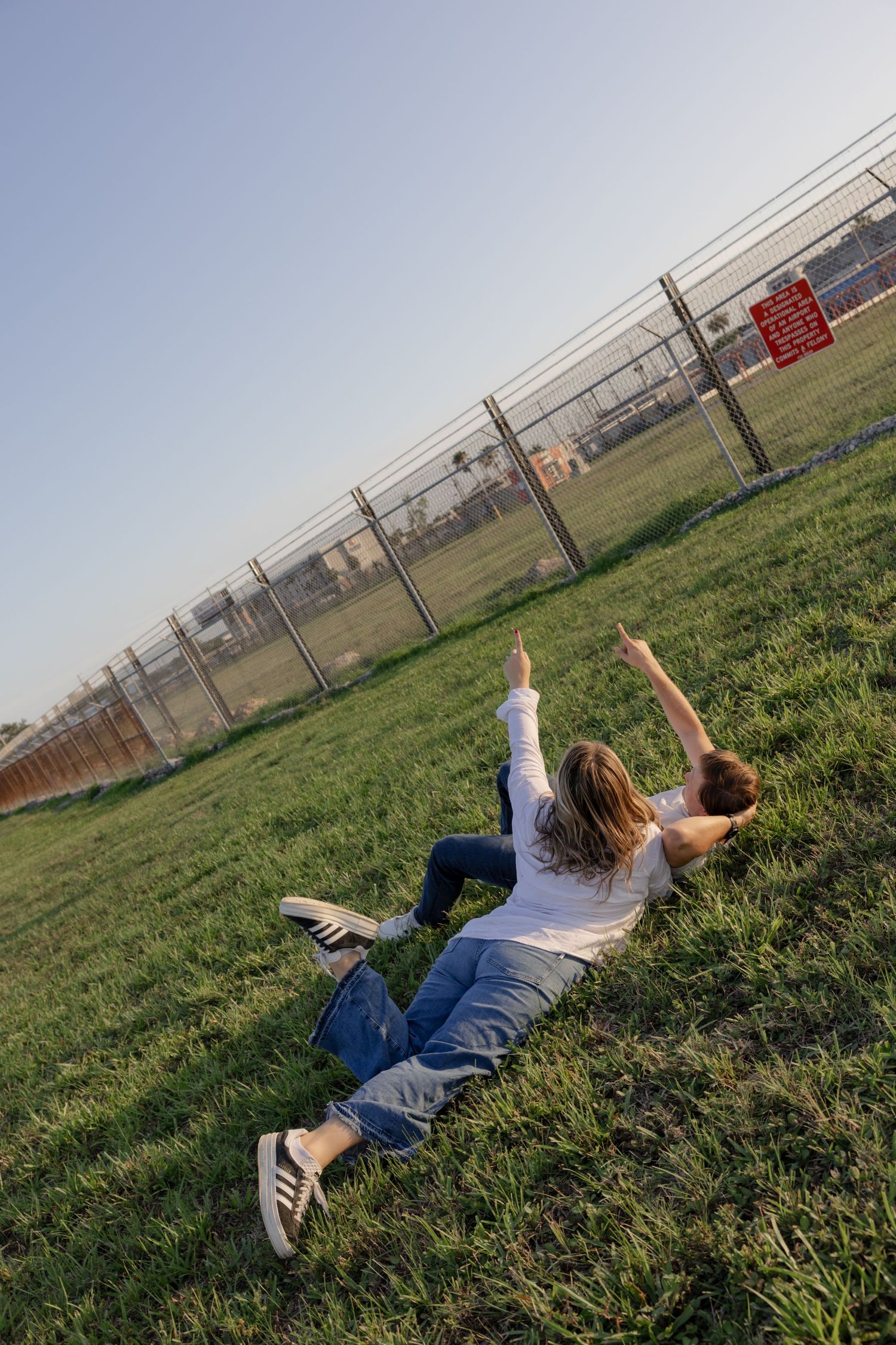 Two people lying on grass watching planes fly overhead near a fence.