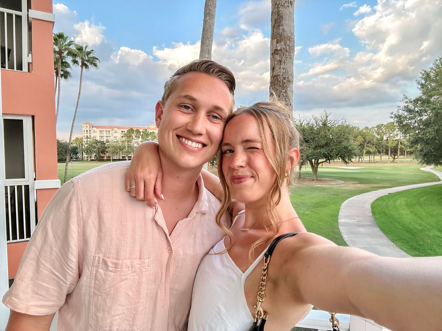 Couple taking selfie photos together outside on a sunny day with palm trees and buildings in background.