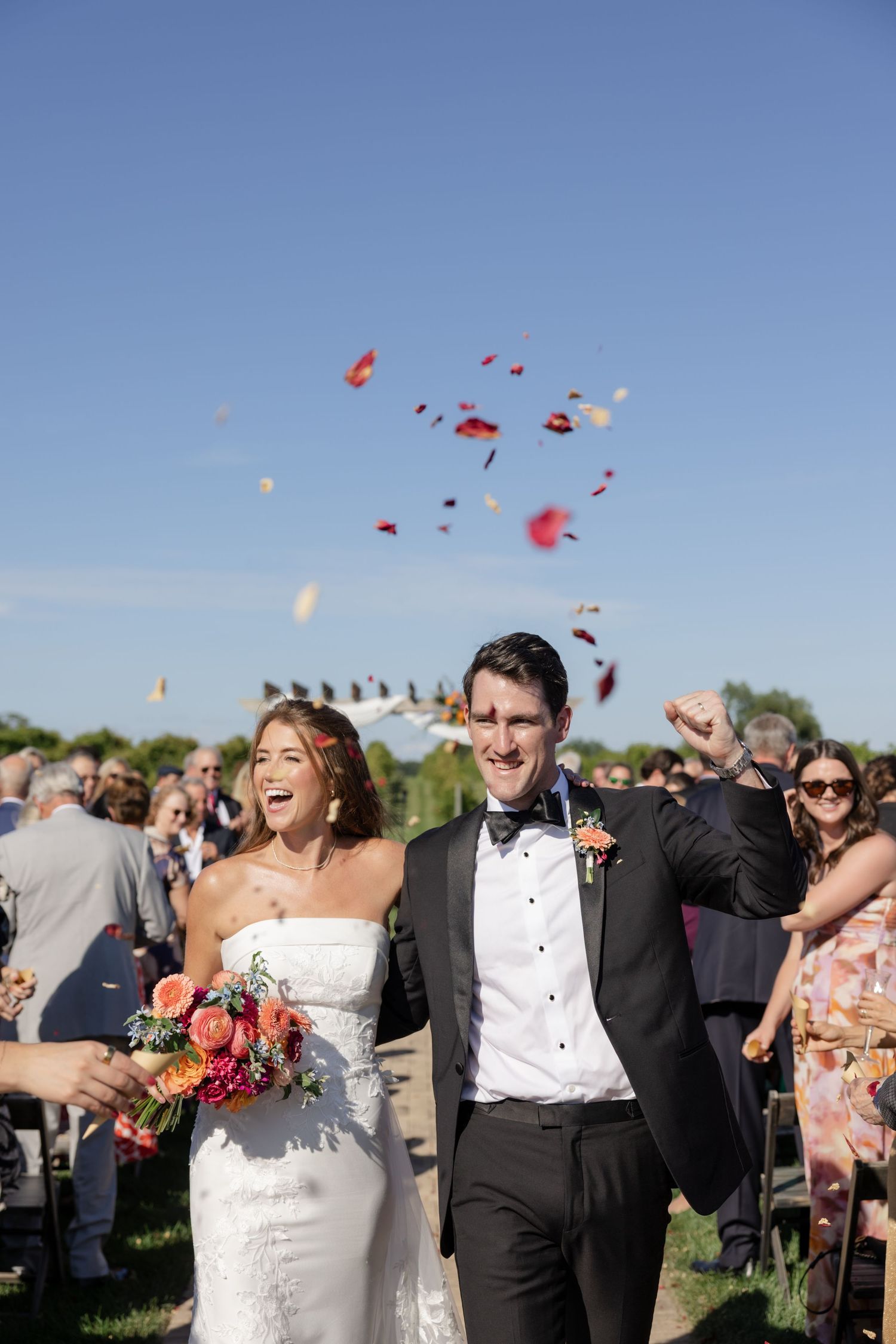 Wedding guests celebrate outdoors as red flower petals are tossed into the bright blue sky.