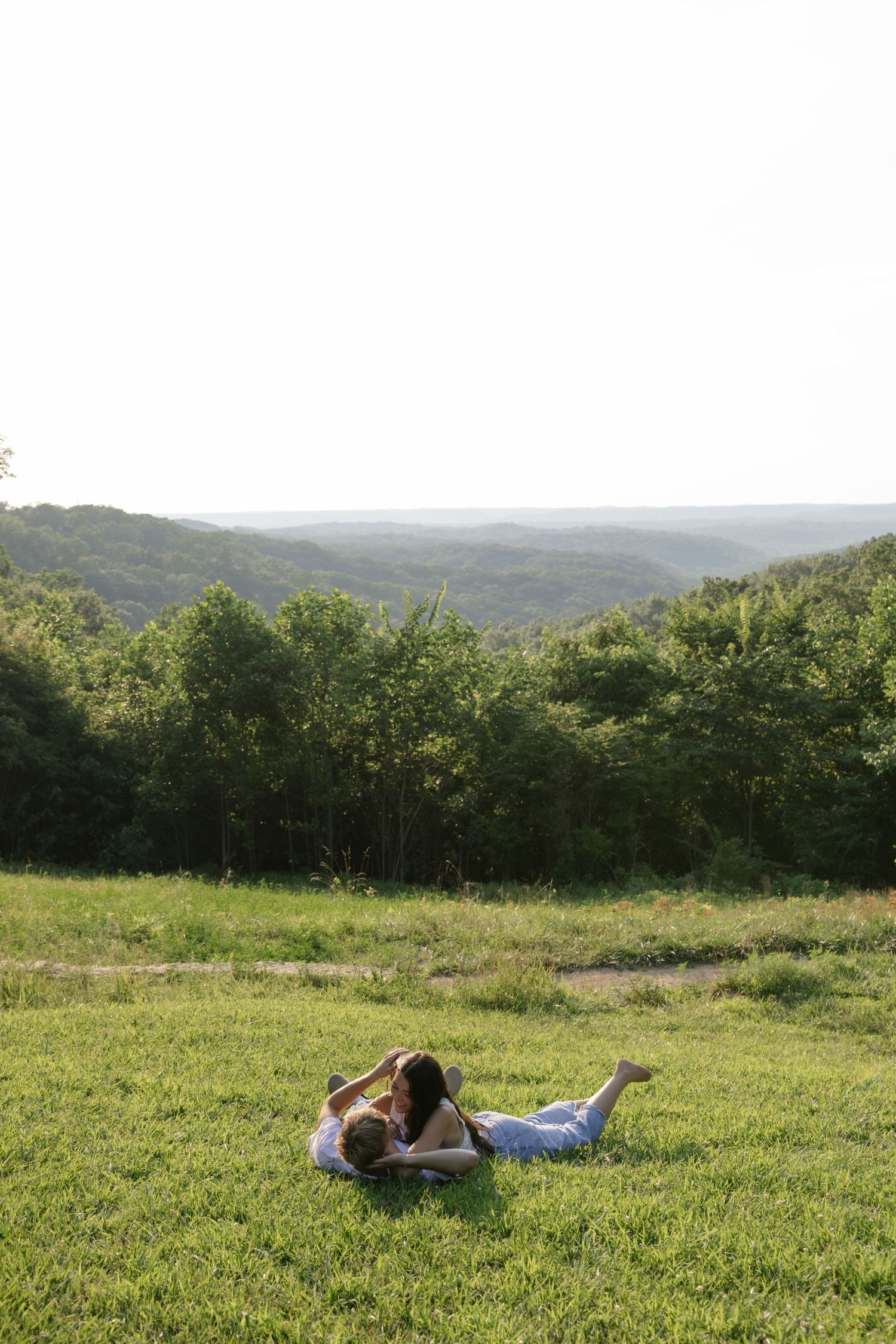 A couple rests on a grassy hillside overlooking a scenic forest valley view.