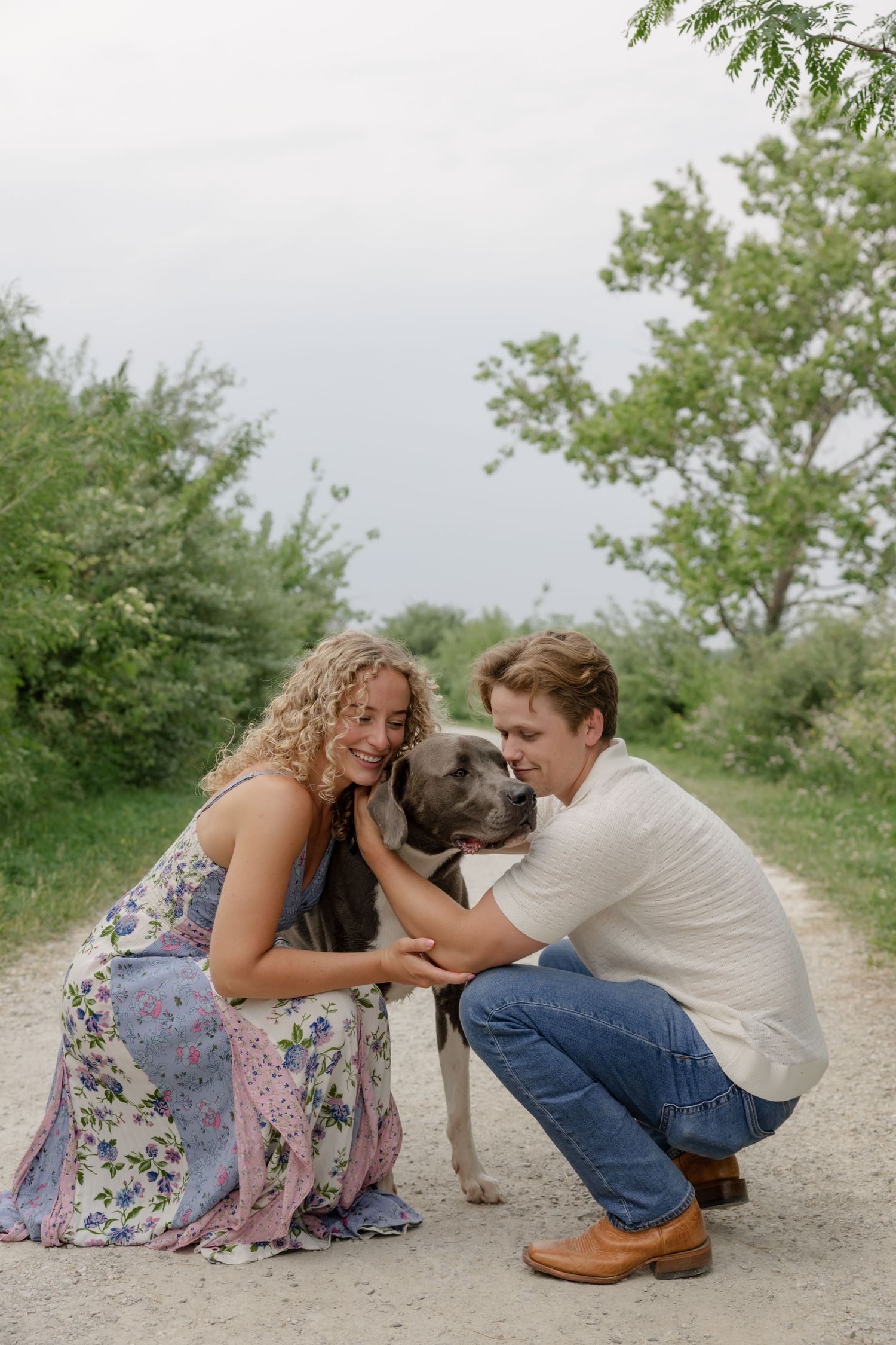 A couple crouches down to pet their happy dog on a nature path surrounded by greenery.