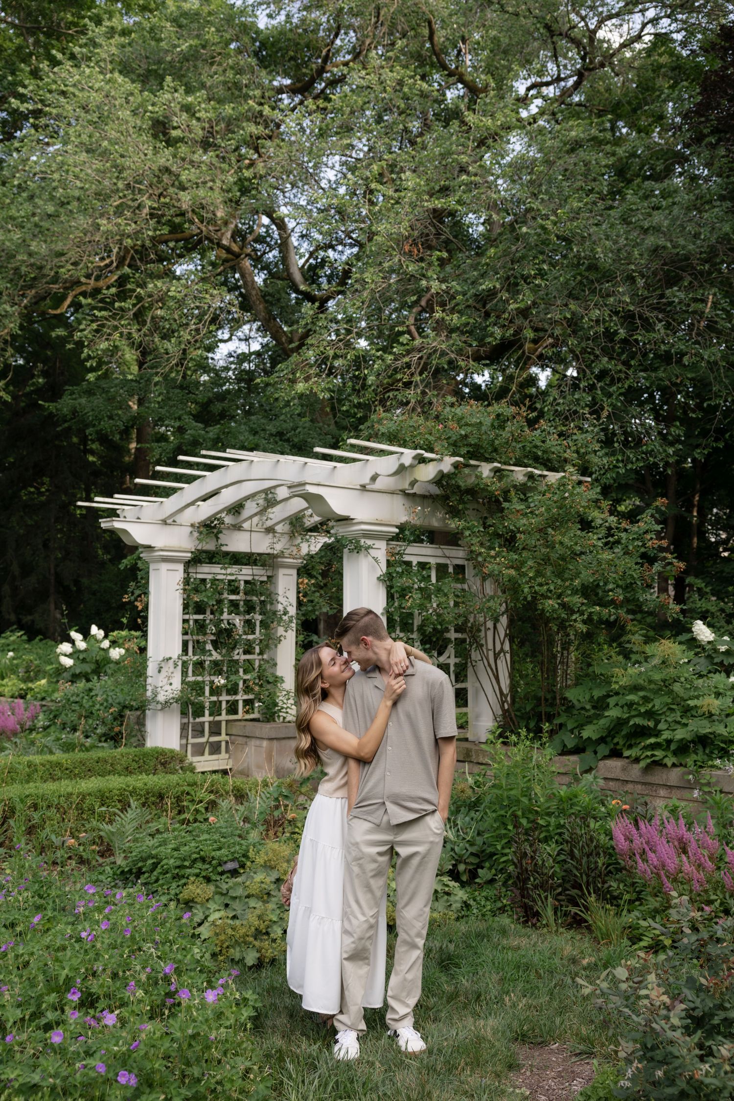 A couple embraces in front of a white garden pergola surrounded by purple flowers and greenery.