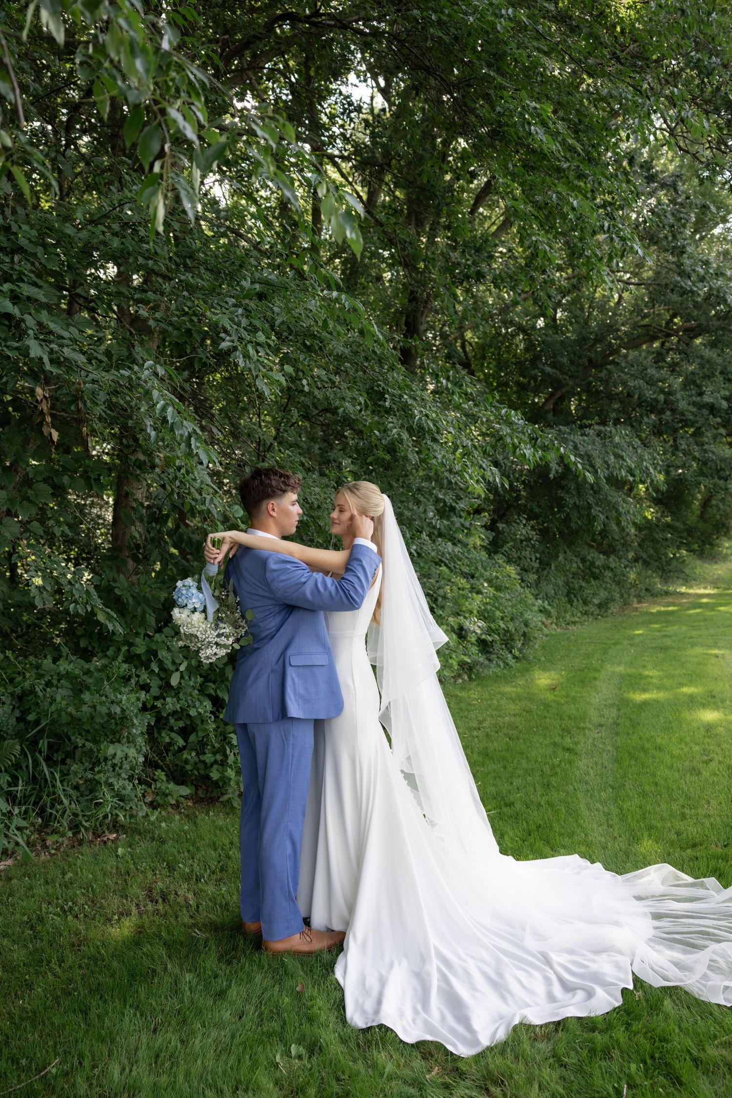 Bride in flowing white dress and groom in blue suit embrace in a garden setting.