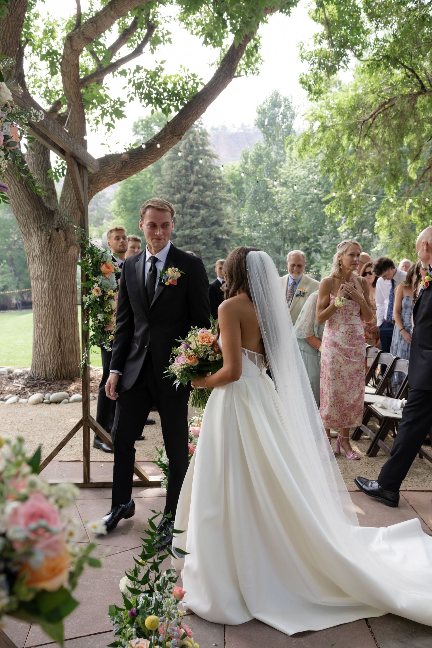 Outdoor wedding ceremony with bride in flowing white dress under tree canopy.
