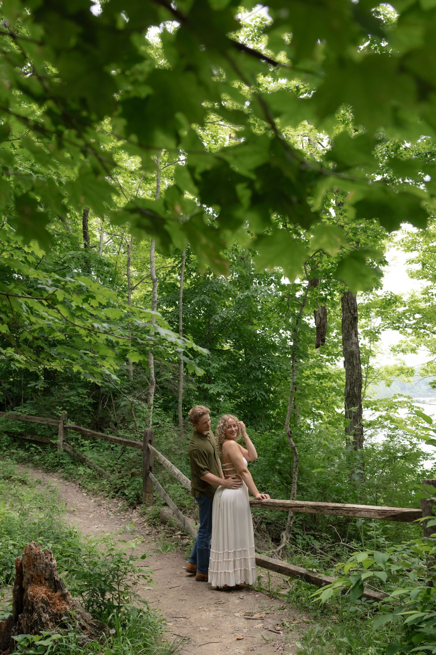 A couple stands together on a wooded hiking trail overlooking water in the distance.