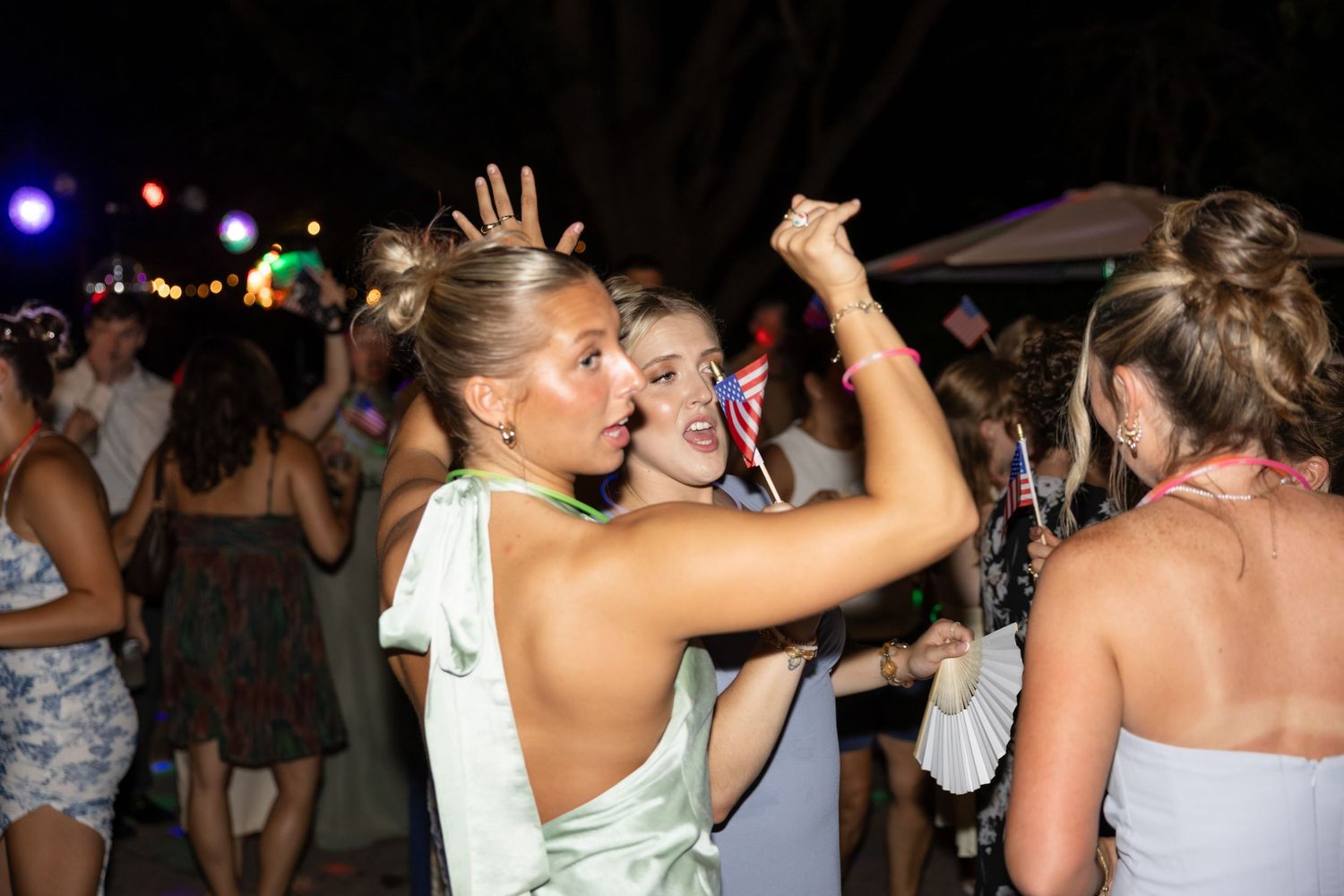 Crowd dancing and celebrating at an outdoor nighttime event with colored lights.