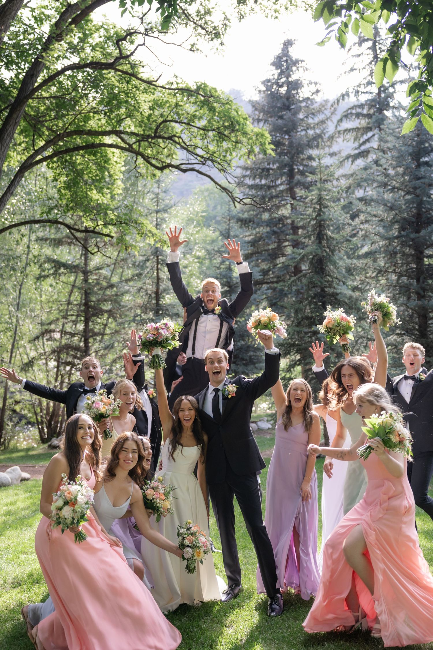 Wedding party celebrating with arms raised in a sunlit garden setting.