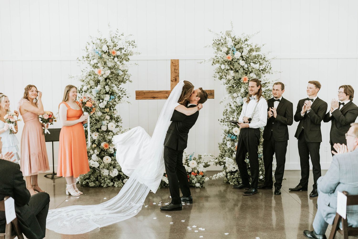 Wedding party lineup in indoor venue with floral arrangements and white chairs.