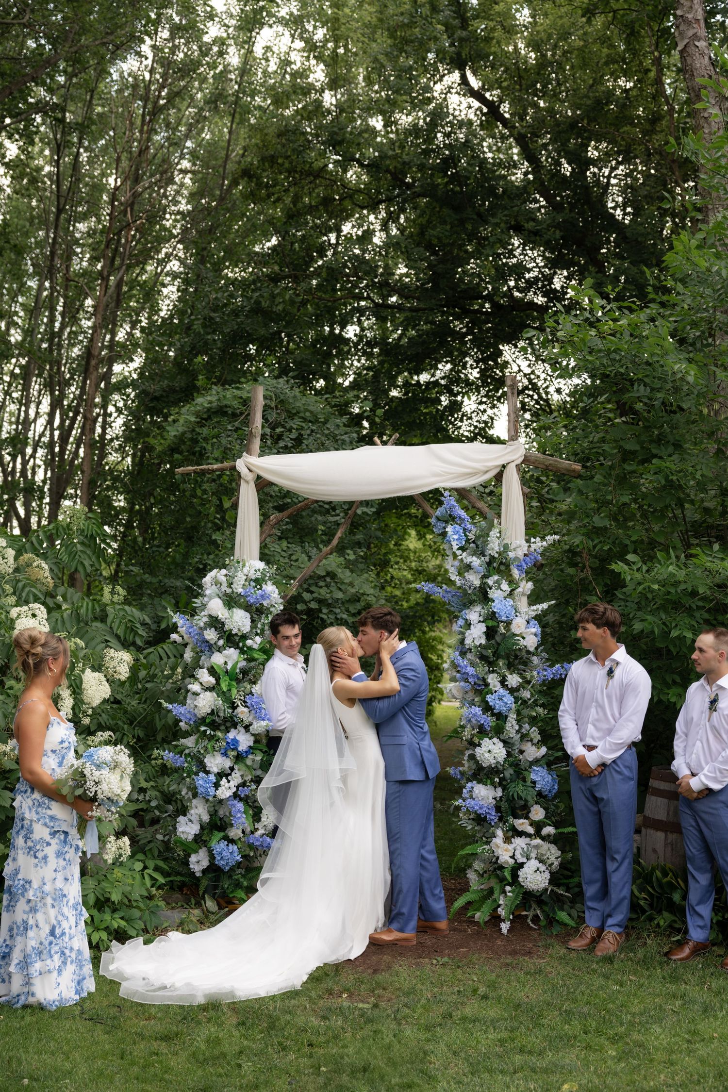 Garden wedding ceremony under trees with blue and white floral decorations.