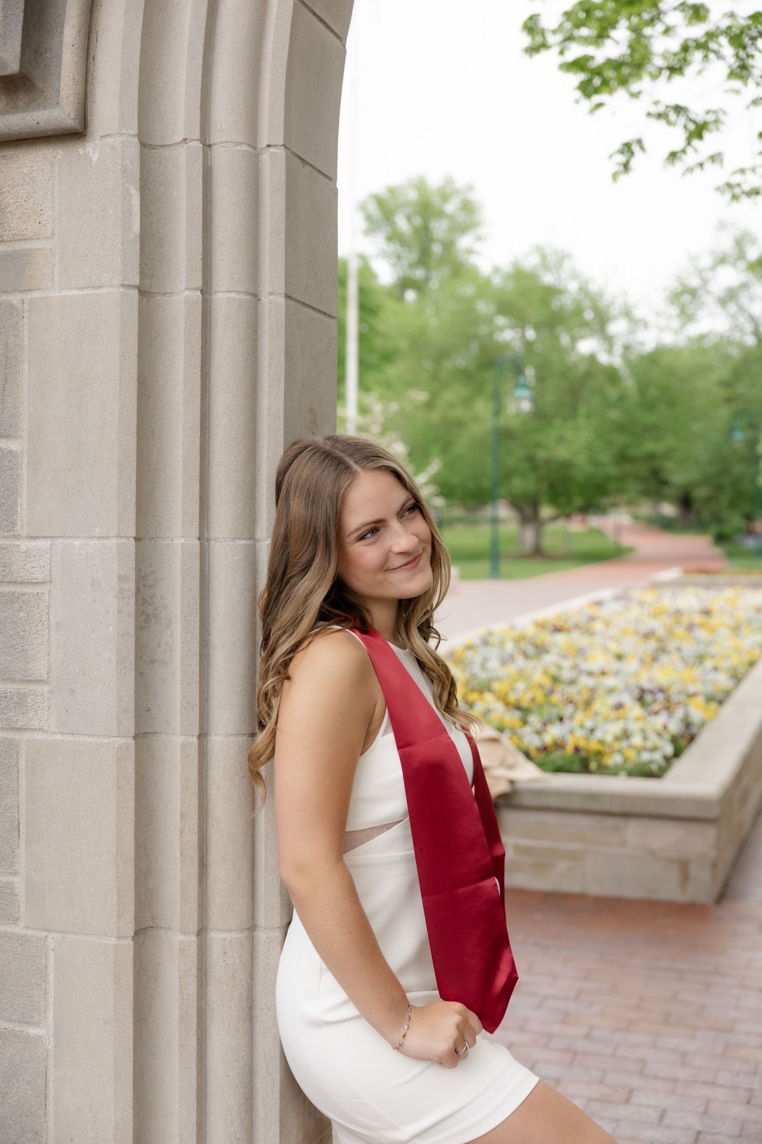 Graduate in white dress and red sash poses against stone column with flowerbeds in background.