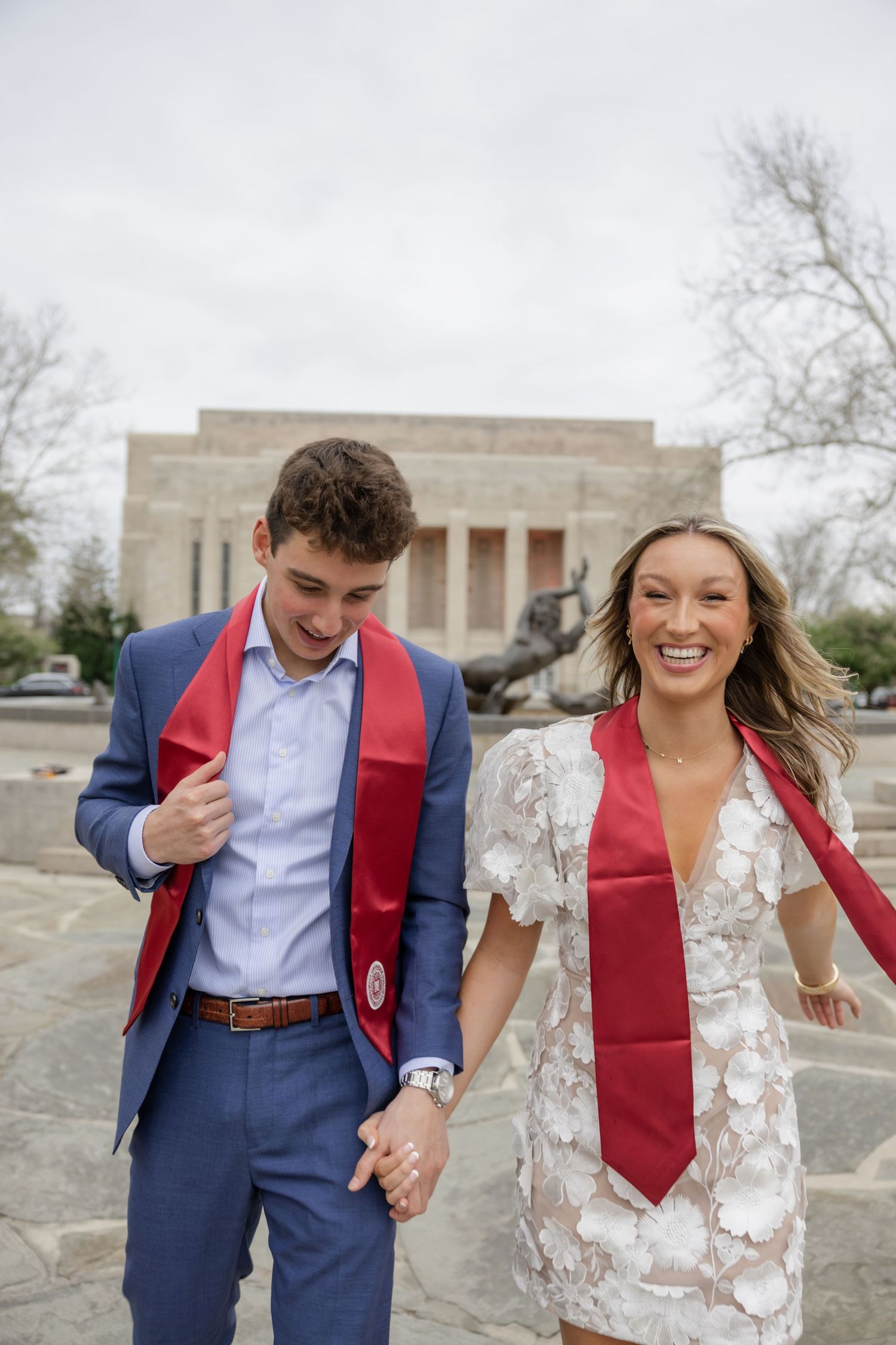 Two people in graduation attire hold hands and laugh while posing outdoors.