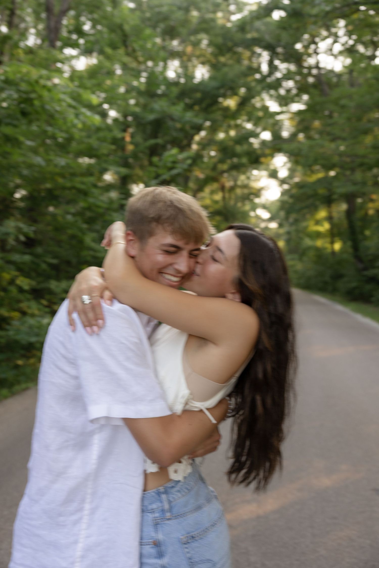A couple in white and denim share playful embraces on a tree-lined path.