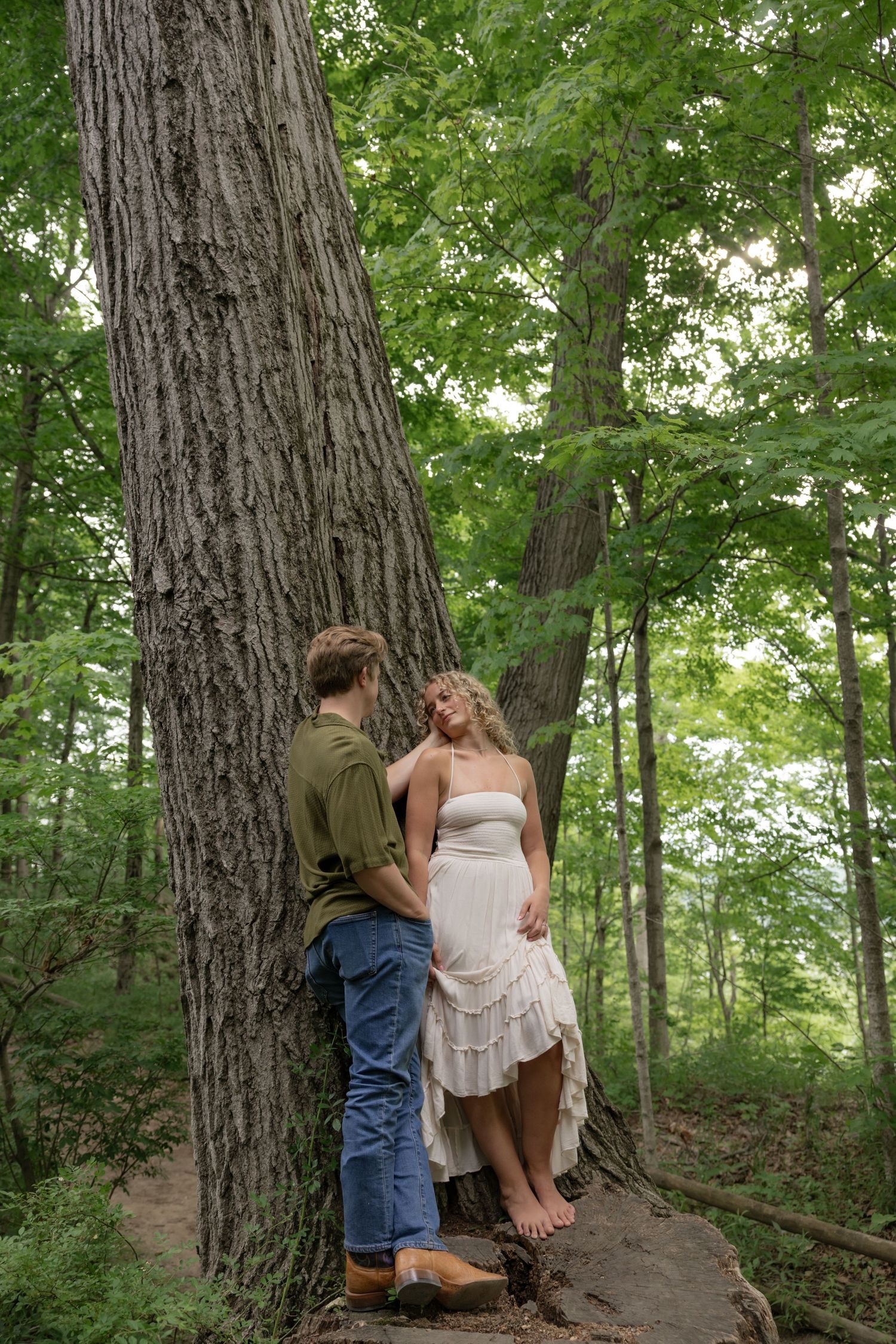 A couple shares a romantic moment against a large tree trunk in a wooded setting.
