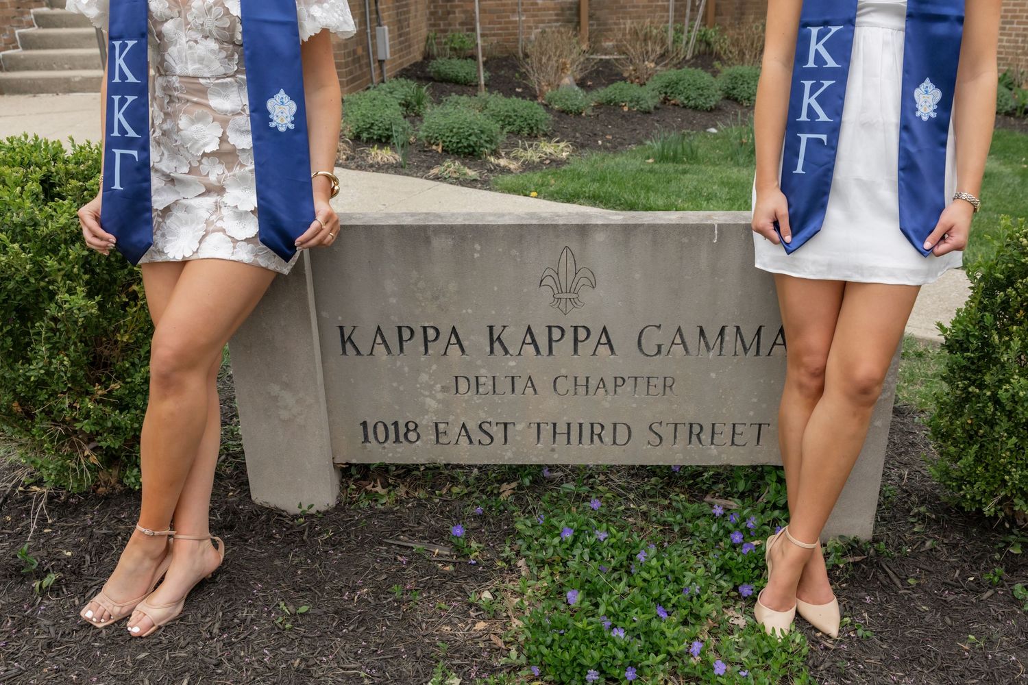 Two pairs of feet stand next to a Kappa Kappa Gamma sorority house sign.