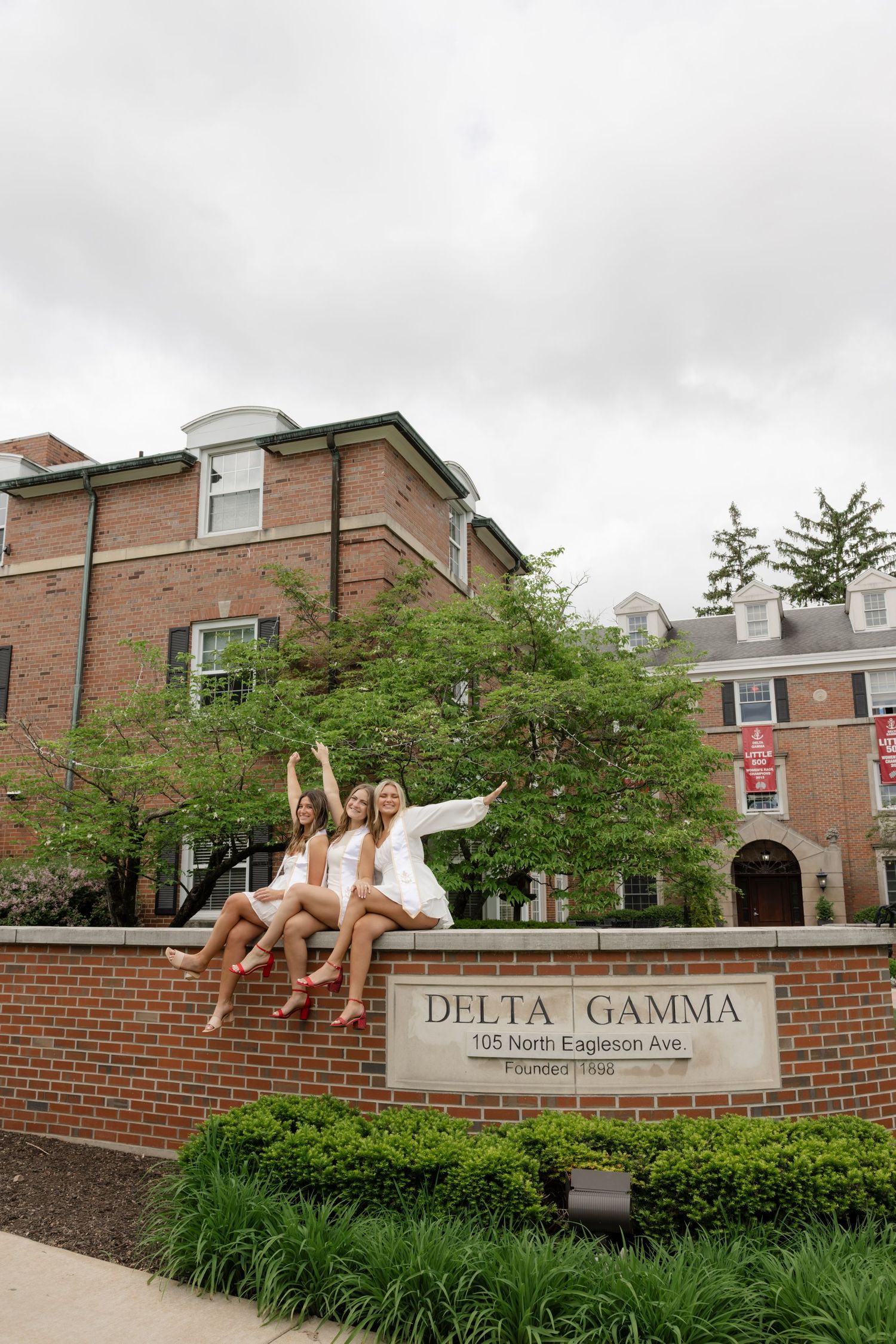 Three graduates sitting on a Delta Gamma sorority house sign in white dresses and red sashes.