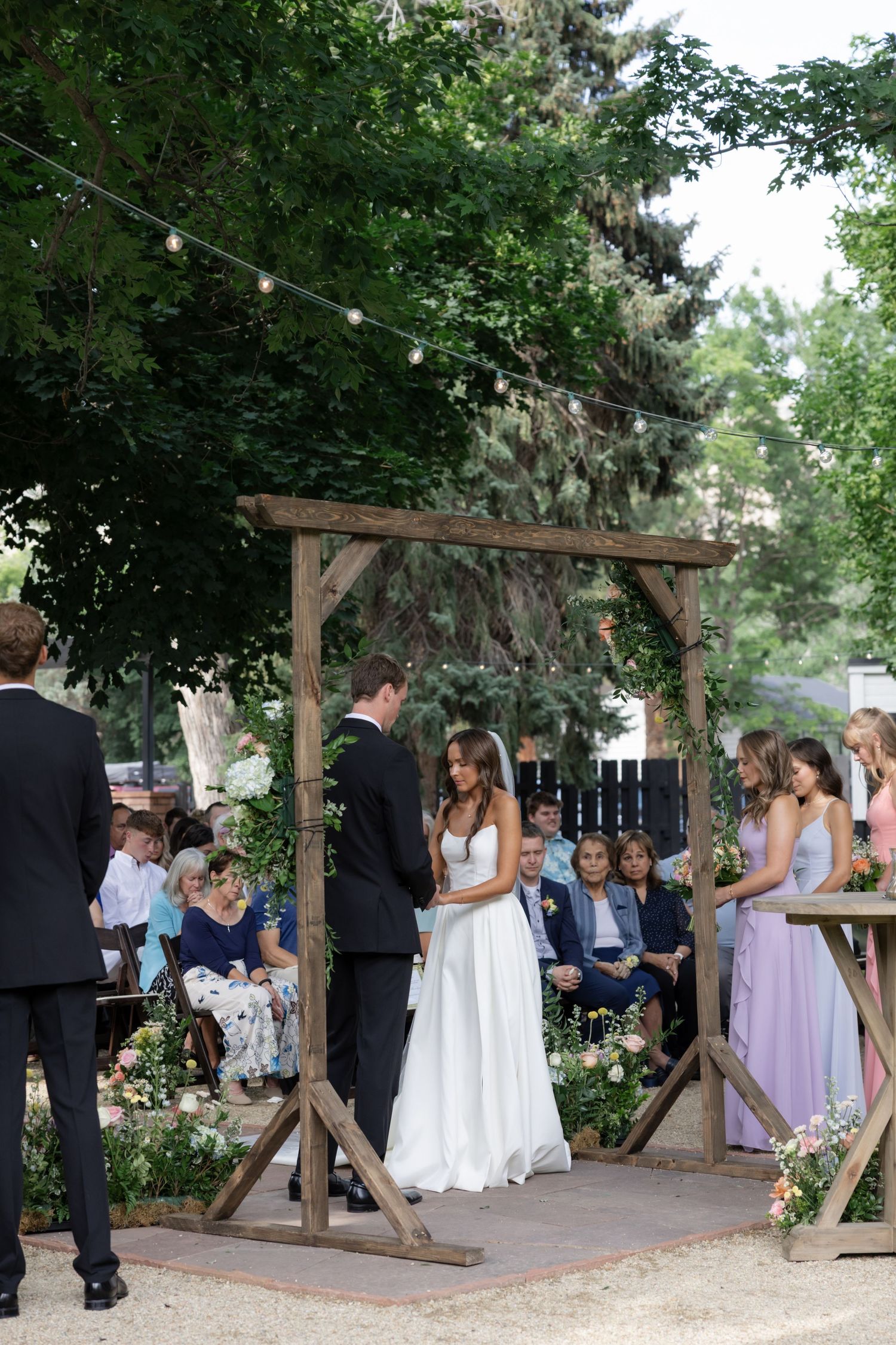 Wedding guests gather under string lights for outdoor garden ceremony.