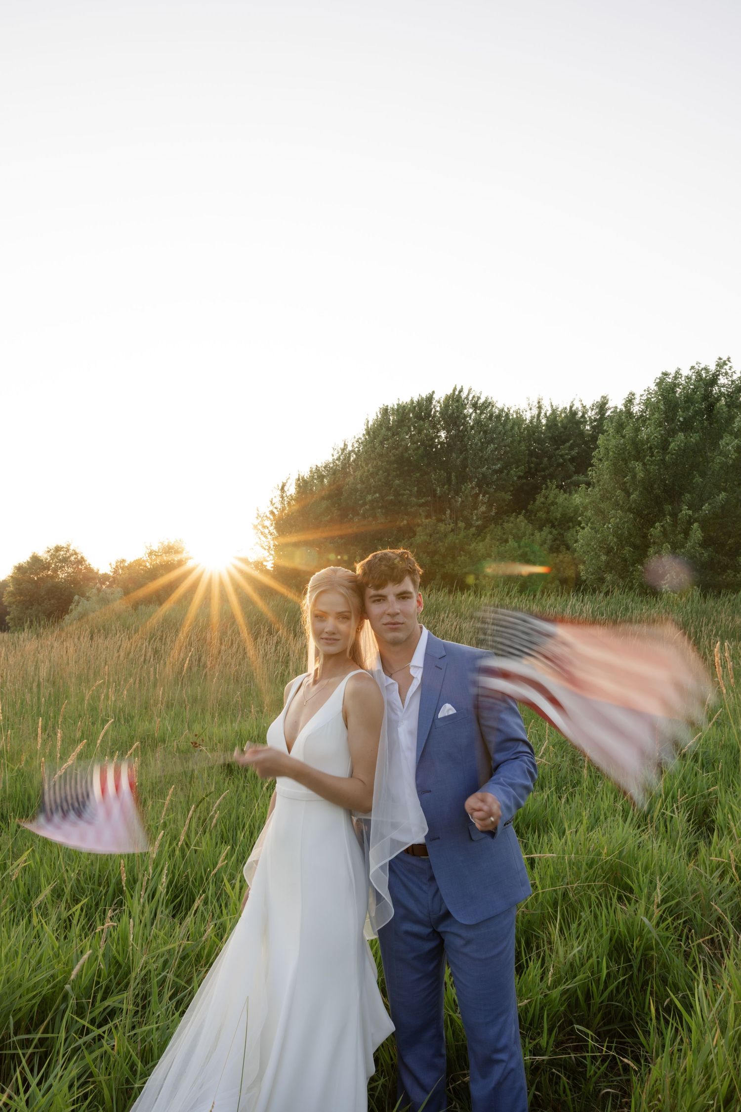 Newlyweds playfully posing with flowing veil during golden hour in a grassy field.
