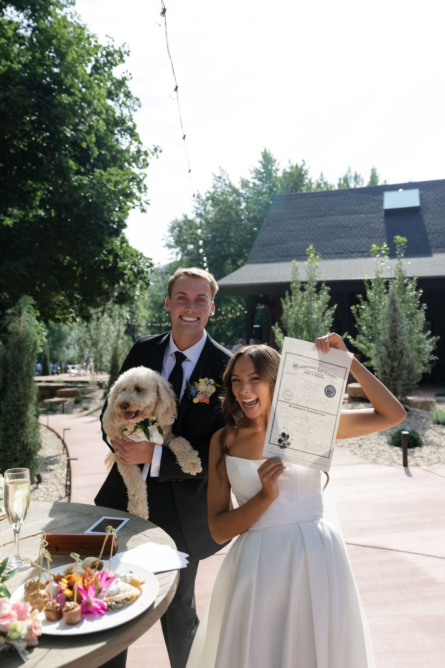Newlyweds holding up their marriage certificate while enjoying a celebratory meal outdoors.