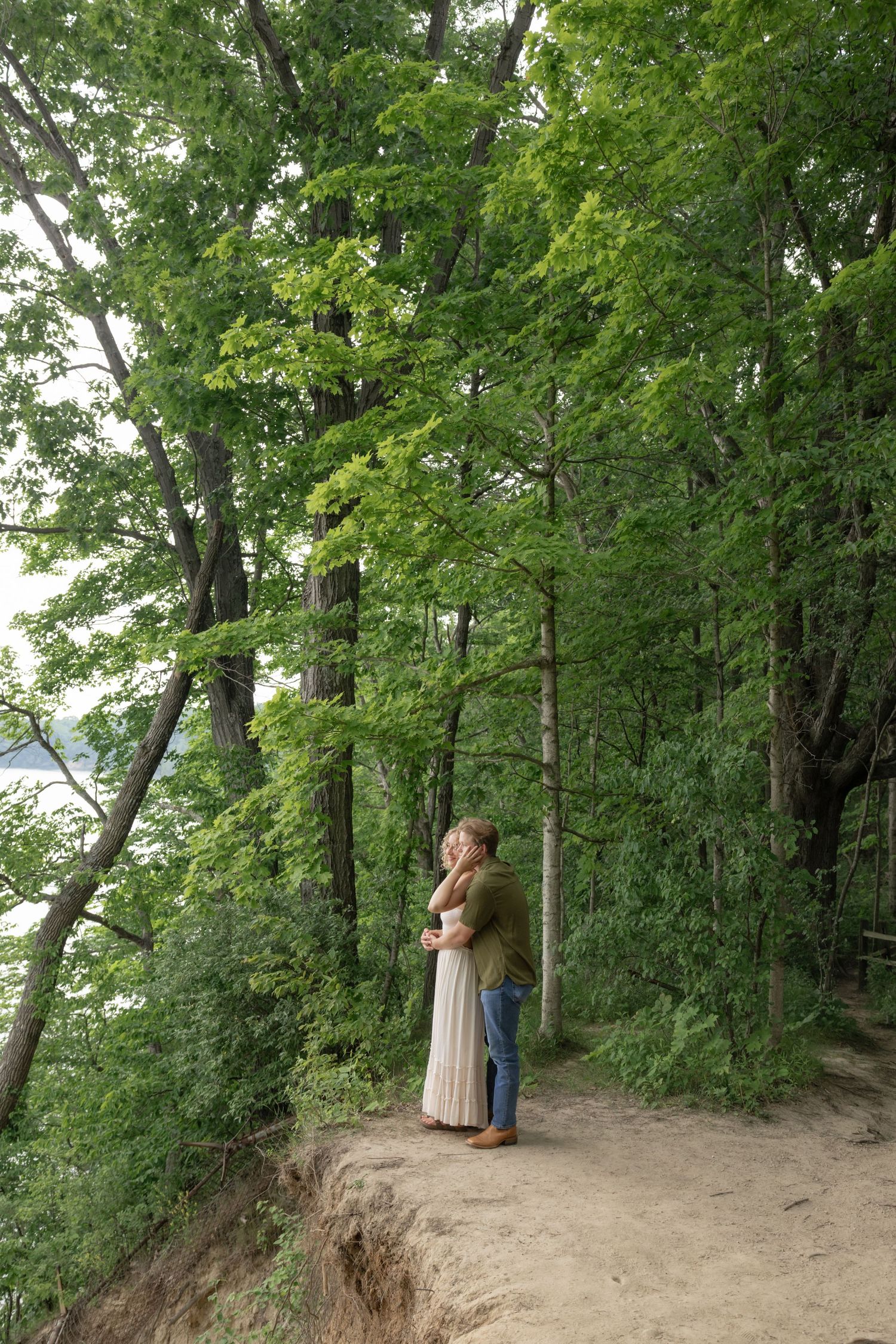A couple stands together on a forested hillside path surrounded by tall trees.