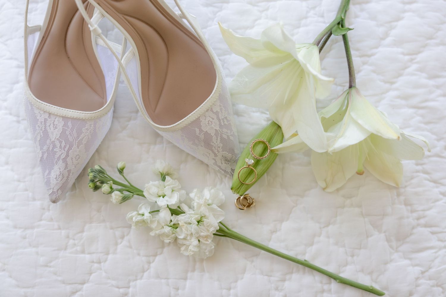 Elegant white bridal shoes and delicate floral bouquet arranged on lace fabric background.