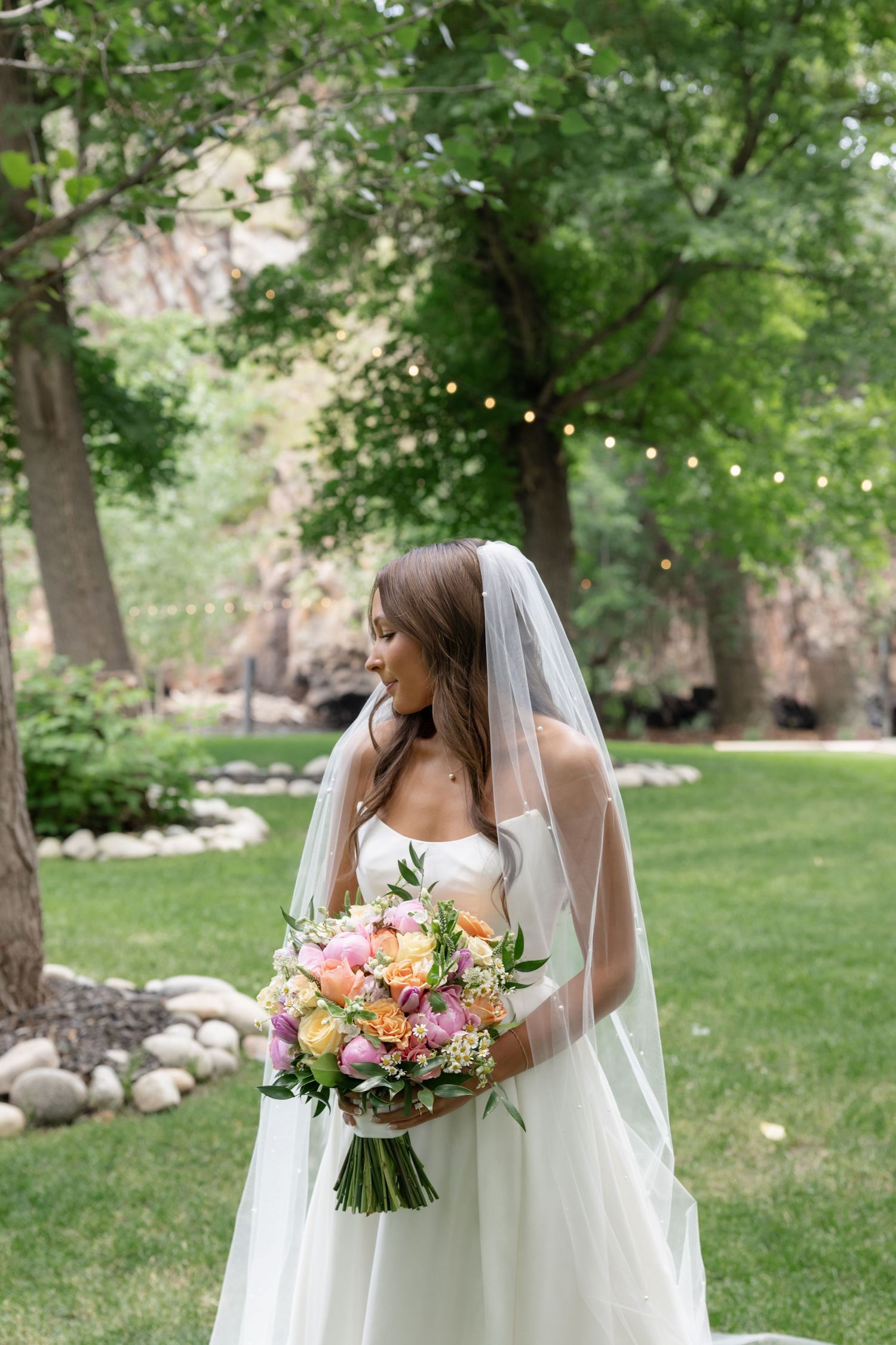 Bride holds pastel floral bouquet against white dress in outdoor garden setting.