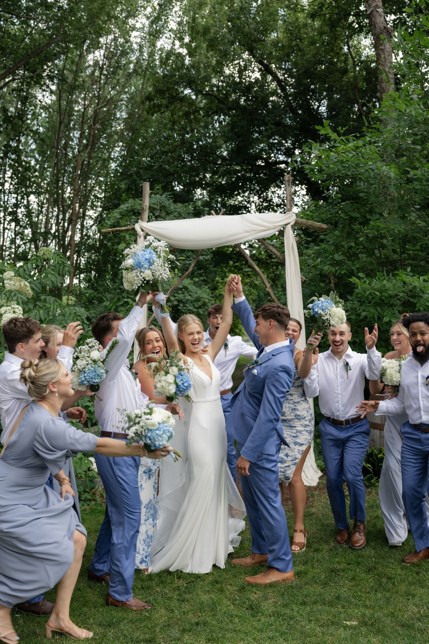 Joyful wedding party celebrating with arms raised under an outdoor ceremony arch.