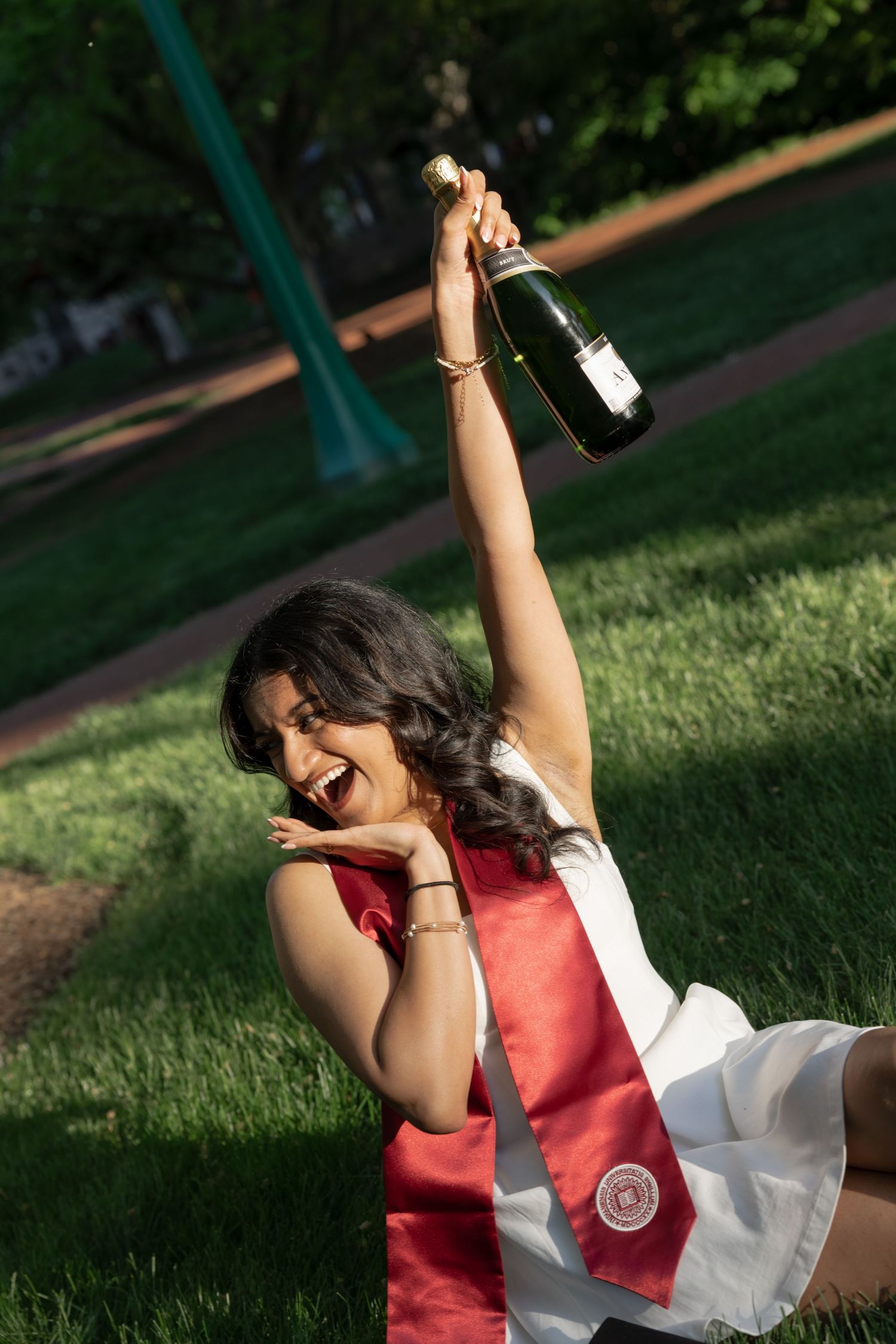 A person in graduation attire celebrates joyfully on grass with raised arms.