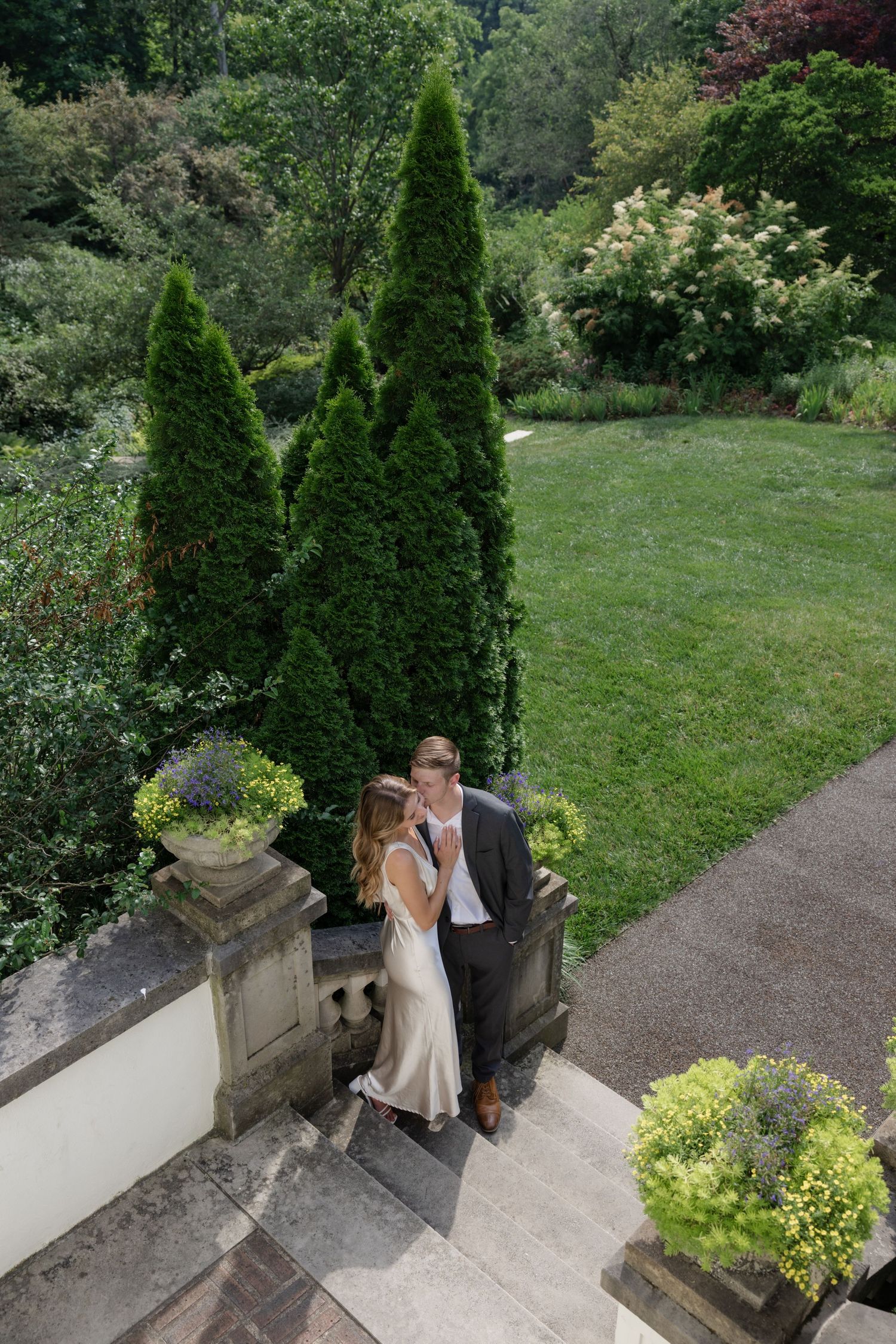 Wedding couple walking down garden path surrounded by manicured evergreen trees.