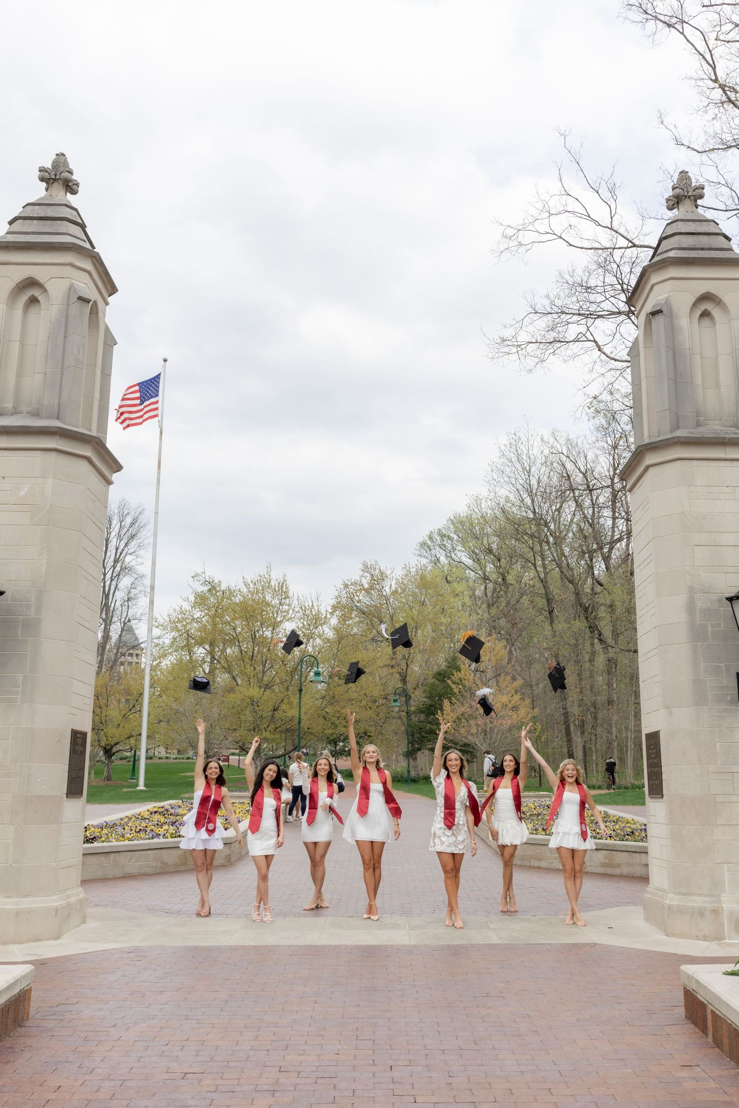 Group of graduates in white dresses throw caps between stone pillars at campus entrance.