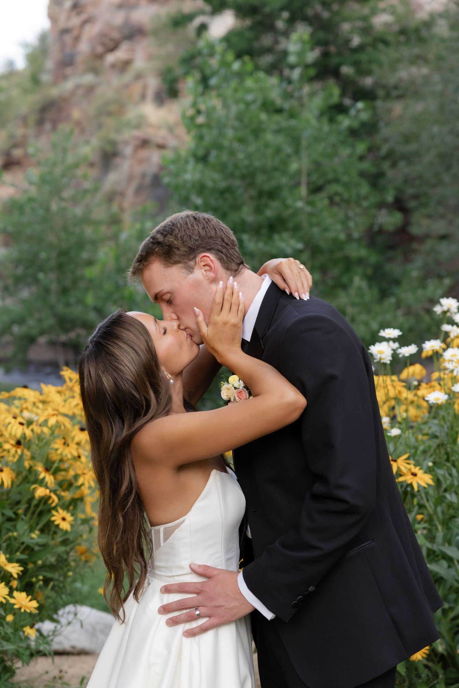 A romantic wedding kiss moment surrounded by blooming yellow flowers and mountain scenery.