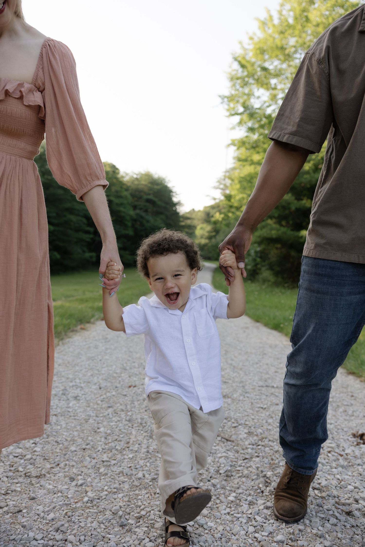 Joyful toddler swings between parents while walking on a gravel path.