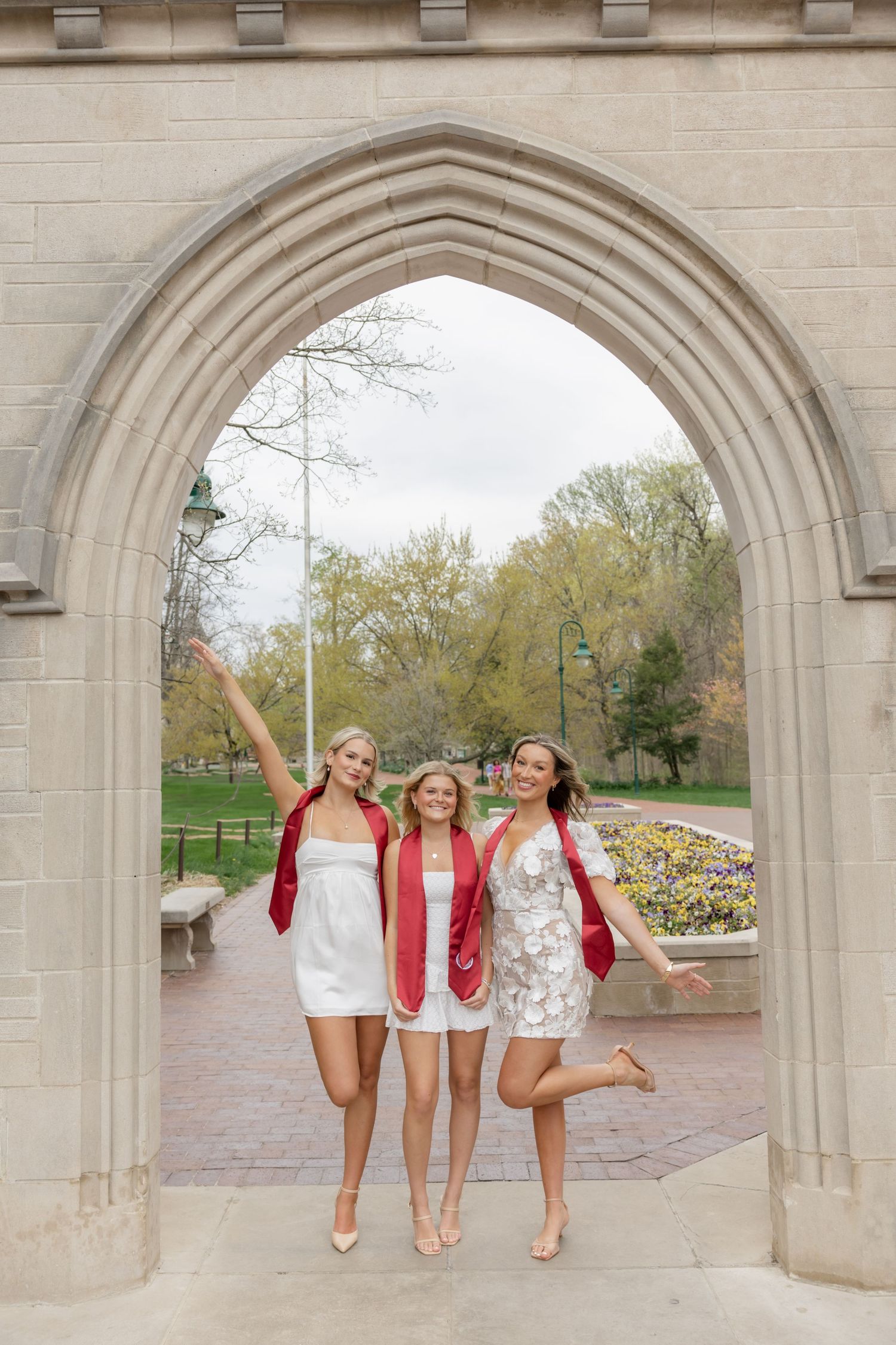 Three graduates in white dresses and red sashes jump joyfully under stone archway.